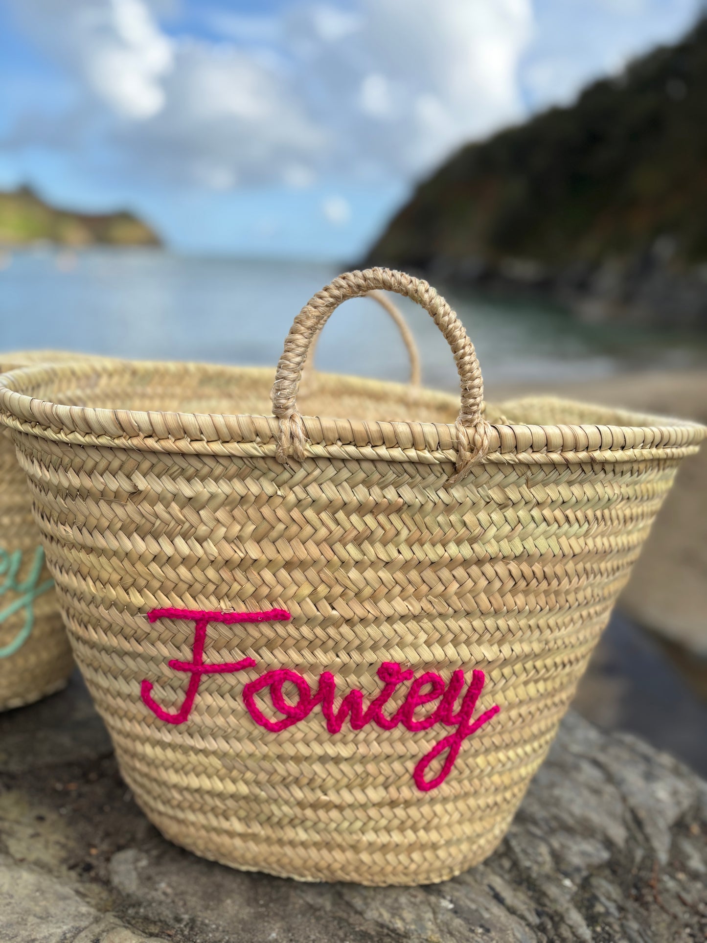 hand-woven straw basket bag with Fowey fuchsia pink embroidery customized beach tote or coastal holiday bag on a Cornish shore