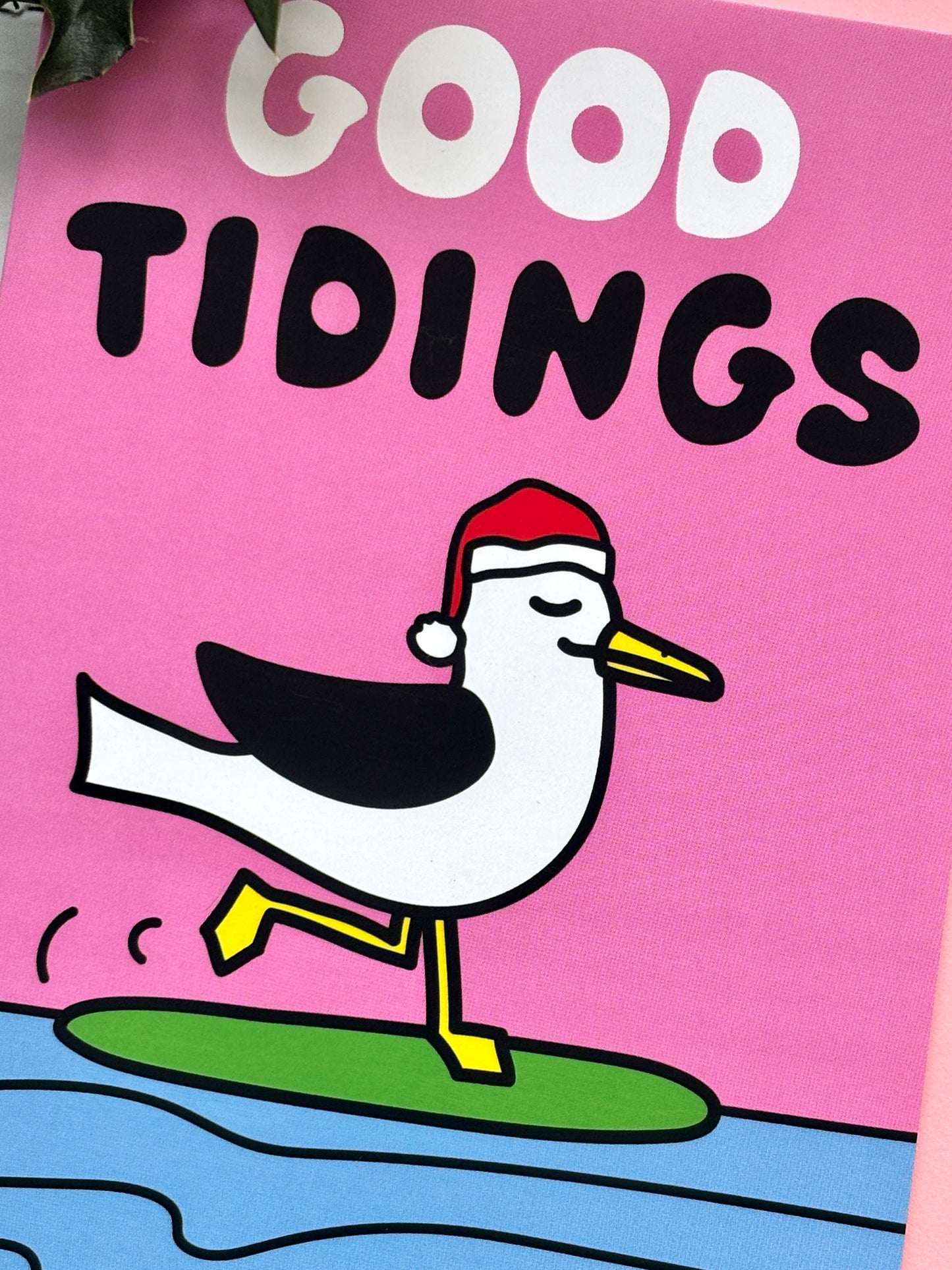 Cartoon seagull in Santa hat with “Good Tidings” text on pink background, standing happily on green surfboard