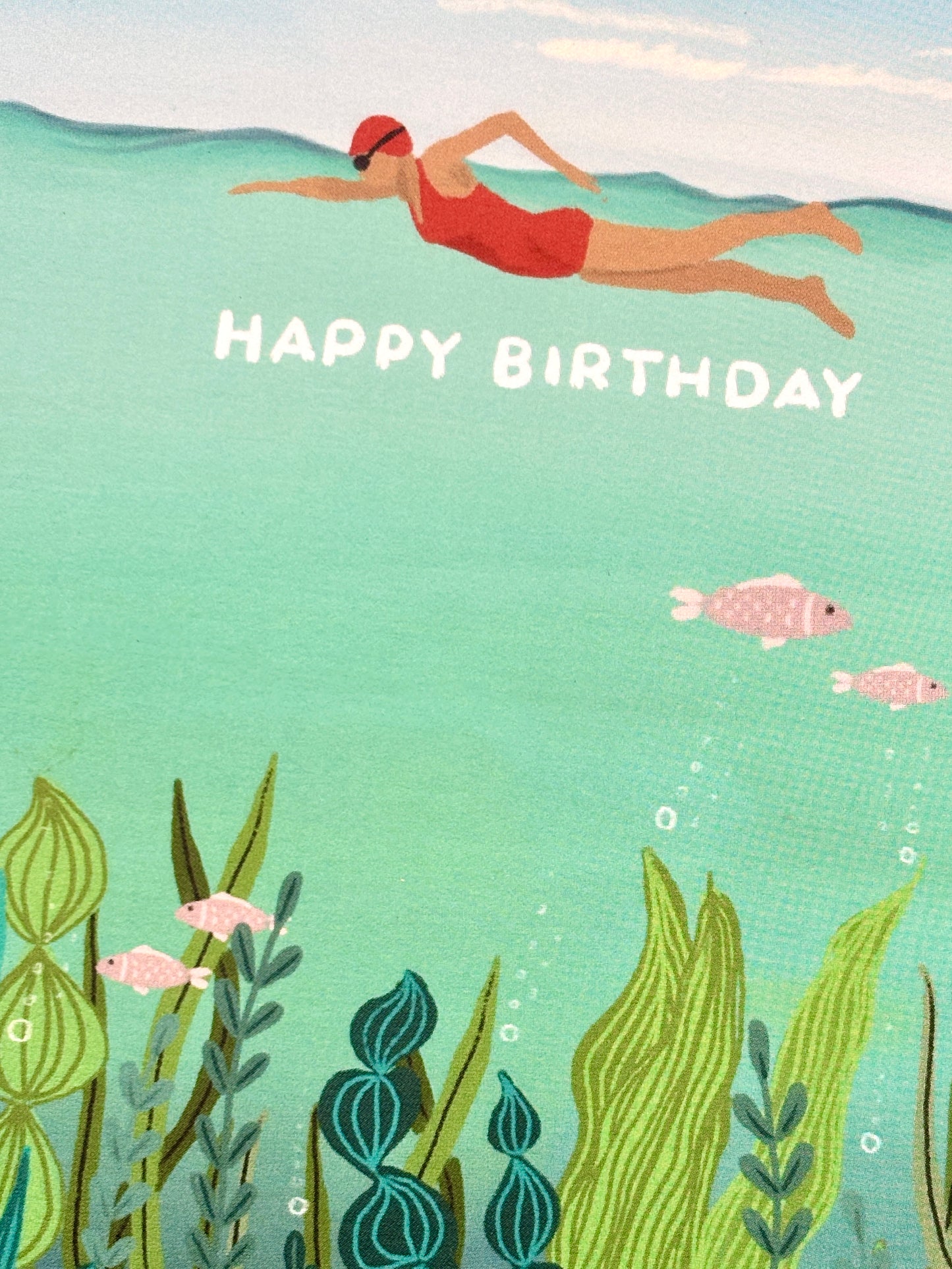 closeup shot of a Happy Birthday card showing an underwater illustration of a swimmer and colorful marine plants and pink fish coastal greeting card for sea swimmers