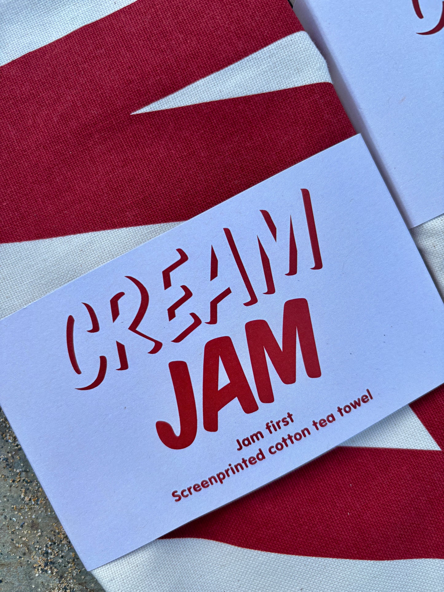 closeup shot of the "CREAM JAM" screenprinted cotton tea towel packaging, highlighting the "Jam first" text funny kitchen linen and Cornwall foodie gift