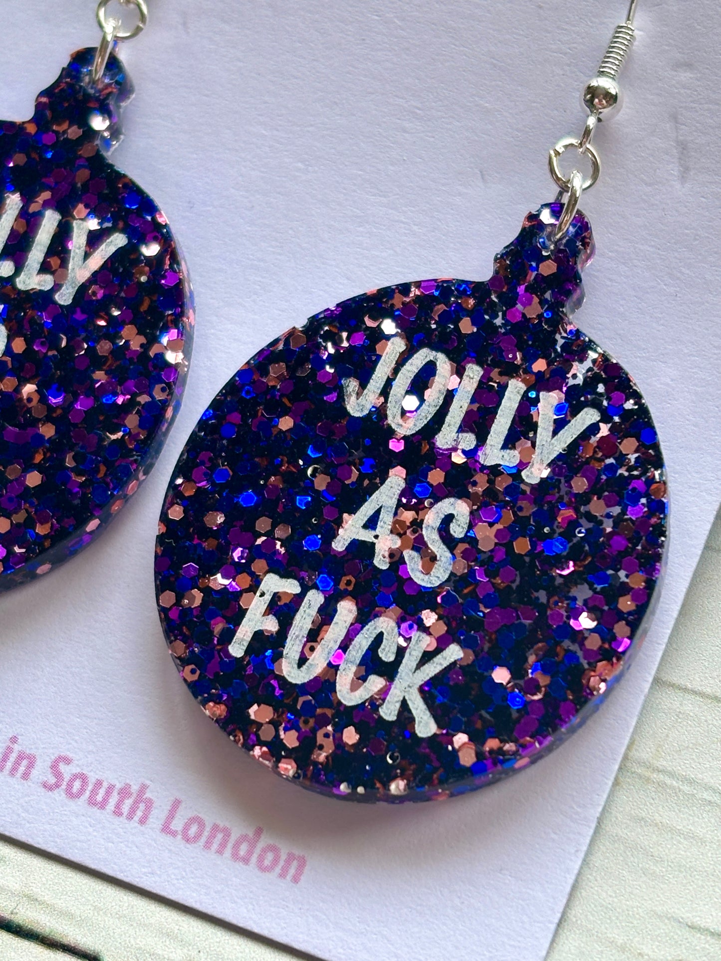 closeup shot of novelty acrylic earring glitter bauble shape with bold, humorous Christmas text unique adult holiday accessory