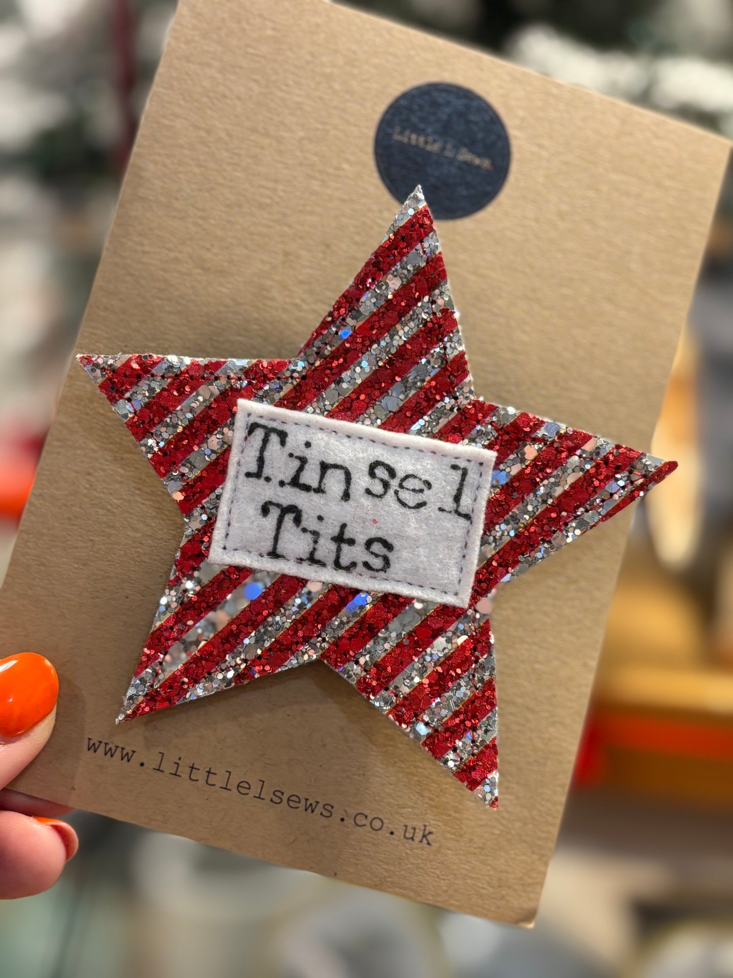 Star-shaped glitter badge with 'Tinsel Tits' label on a brown card, held by a hand with orange nail polish.