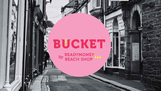 the bright pink logo of Bucket by Readymoney Beach Shop against a black and white photo of Fowey's Fore Street