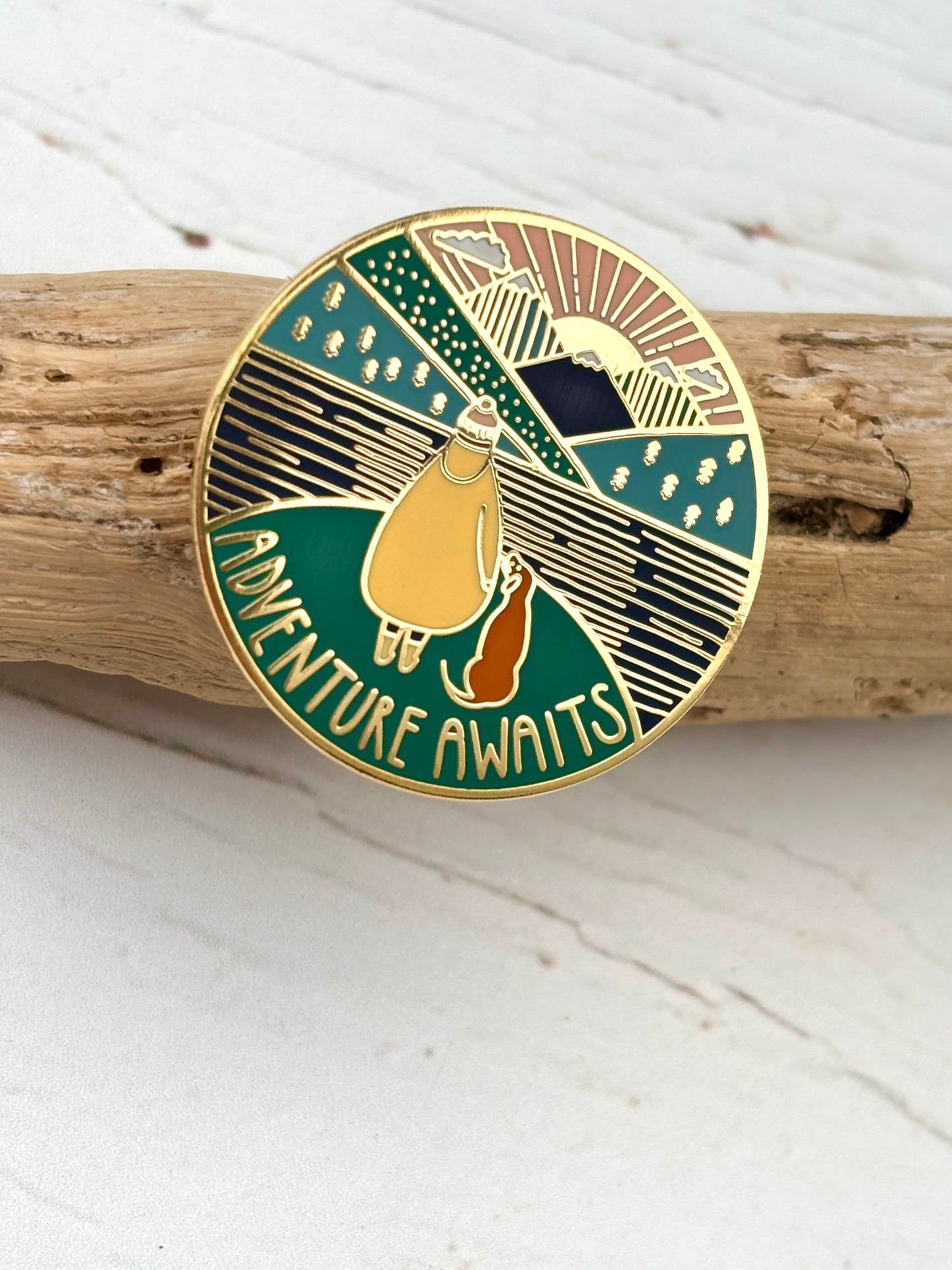 gold enamel hiking pin on driftwood, adventure awaits text, walker with dog, layered mountain landscape design