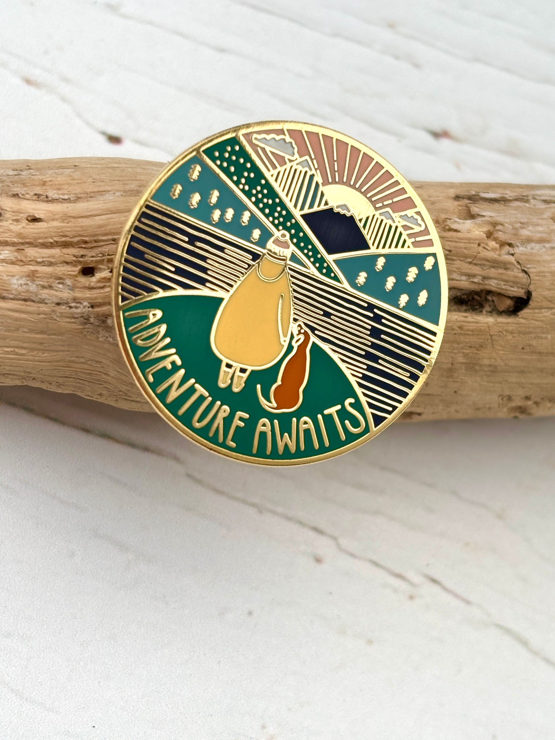gold enamel hiking pin on driftwood, adventure awaits text, walker with dog, layered mountain landscape design
