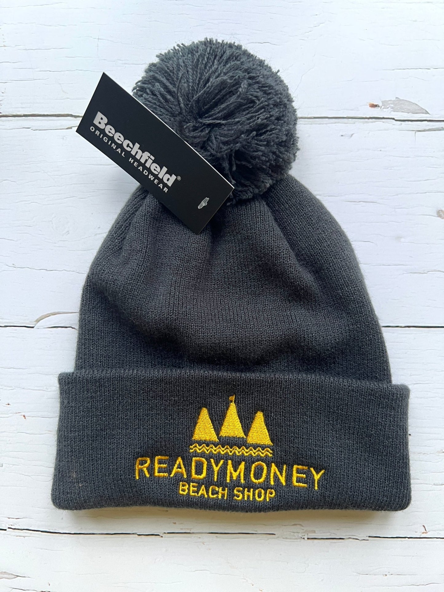 dark grey kids’ beanie with pom-pom, soft knit, and yellow embroidered beach shop logo—warm, durable, and coastal-cool