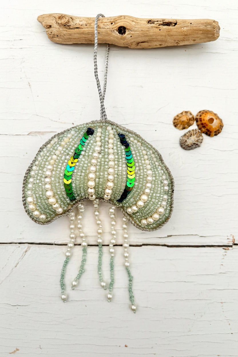 A handmade beaded jellyfish ornament hanging from a driftwood branch on a white wooden surface, with three small seashells placed to the right. The jellyfish is made of pale green and white beads with sequined vertical stripes in green, yellow, and blue, and has pearl-like bead tassels hanging below.
