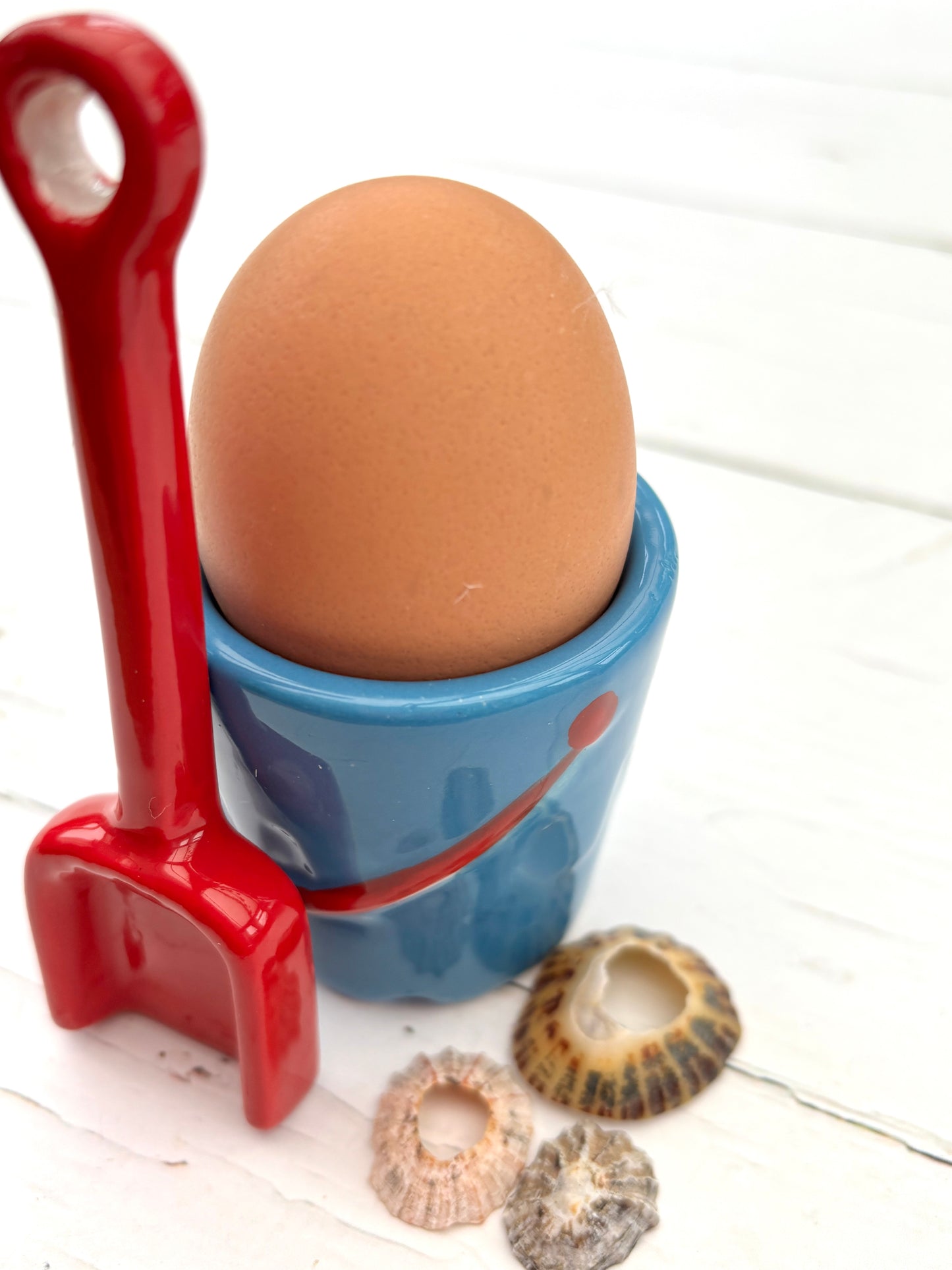 blue ceramic egg cup styled like a beach bucket with red spoon and seashells—fun coastal breakfast table accent