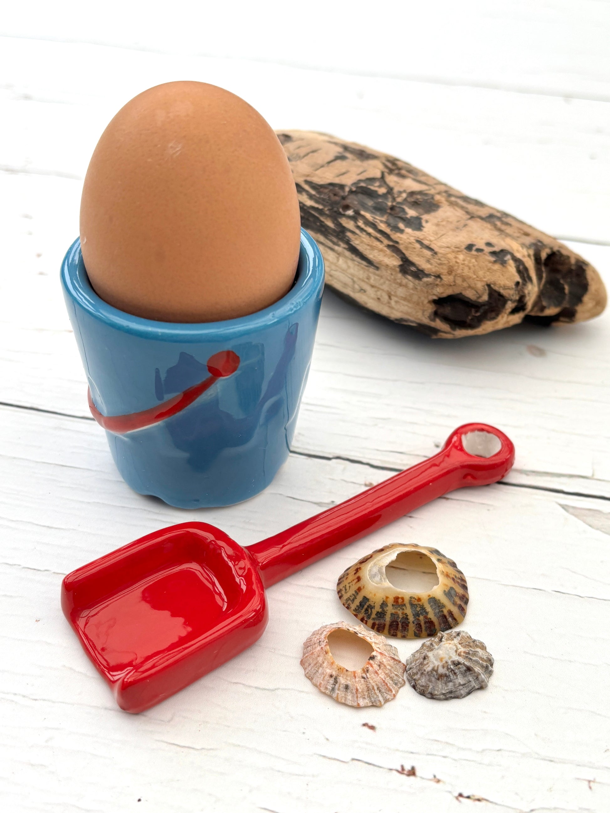 coastal-themed ceramic egg cup with smiley face, red spoon, seashells, and driftwood—playful beach breakfast decor