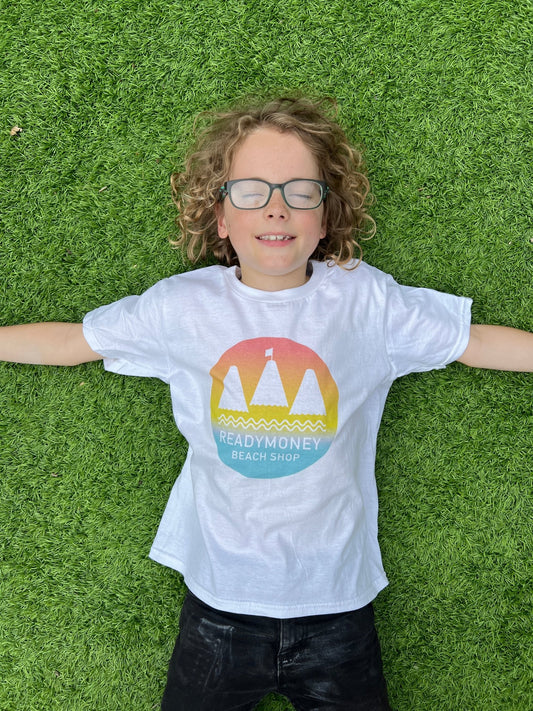 child in white Readymoney Beach Shop T-shirt with colorful sunset logo, lying on grass kids summer apparel, beach souvenir, graphic tee