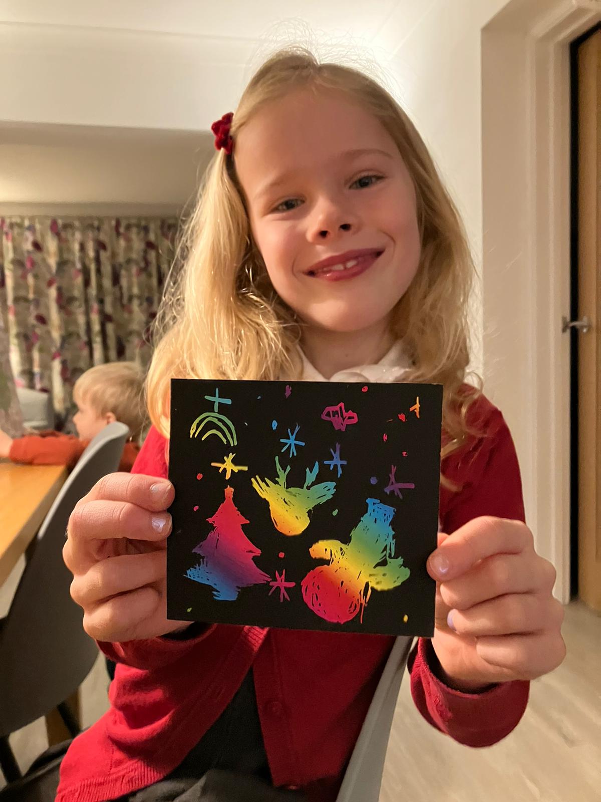 smiling child holding rainbow Christmas scratch art reindeer, tree, and snowman designs kids' holiday craft, creative activity, fun art
