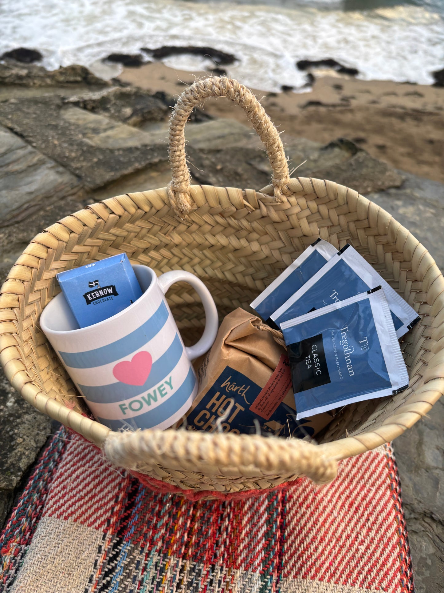 Fowey straw basket on a tartan blanket by the sea contains Kernow chocolate, Fowey mug, and Tregothnan tea luxury coastal gift