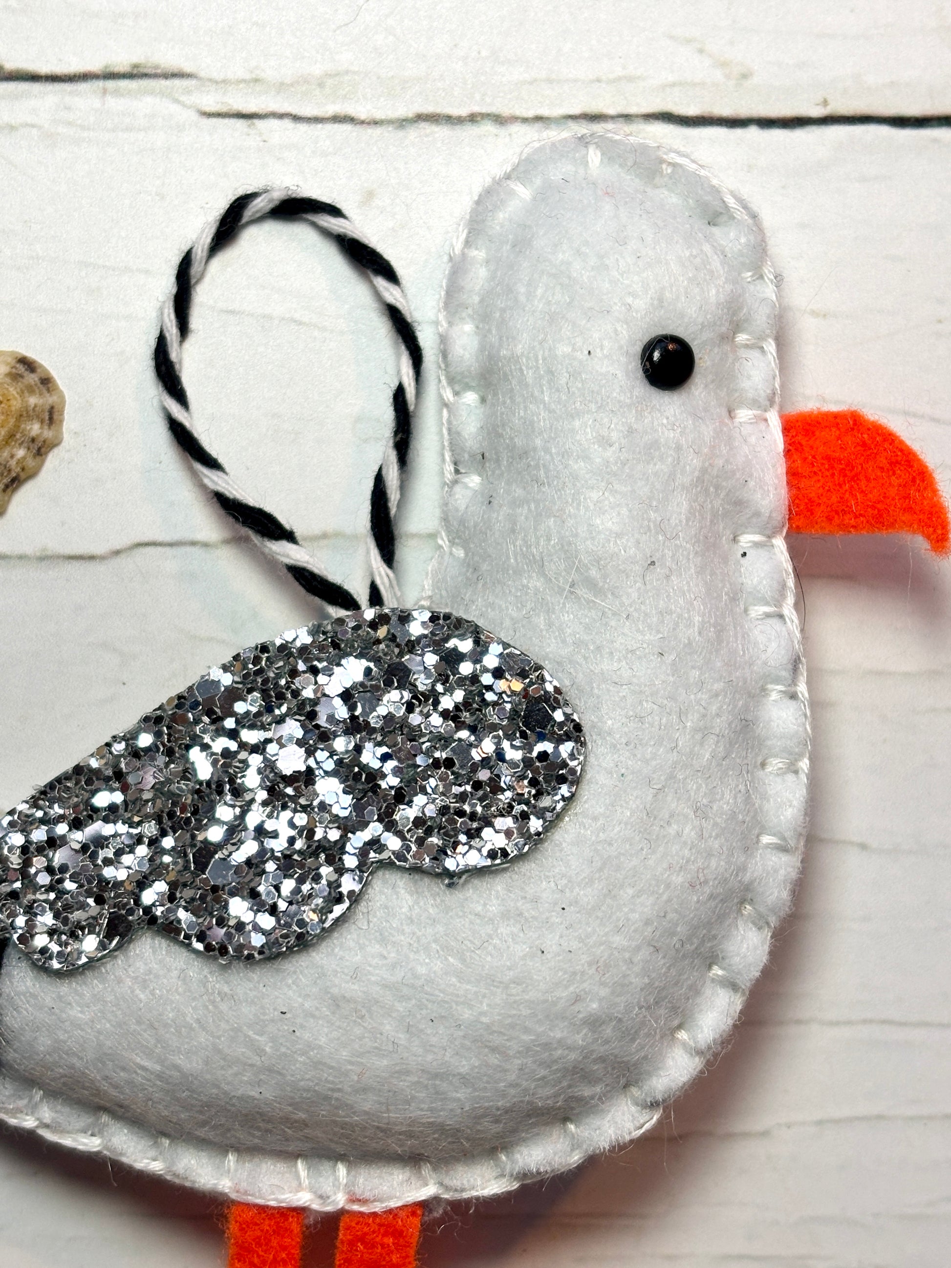 closeup shot of felt seagull ornament handmade coastal decor with glitter wing, orange beak, and nautical striped loop