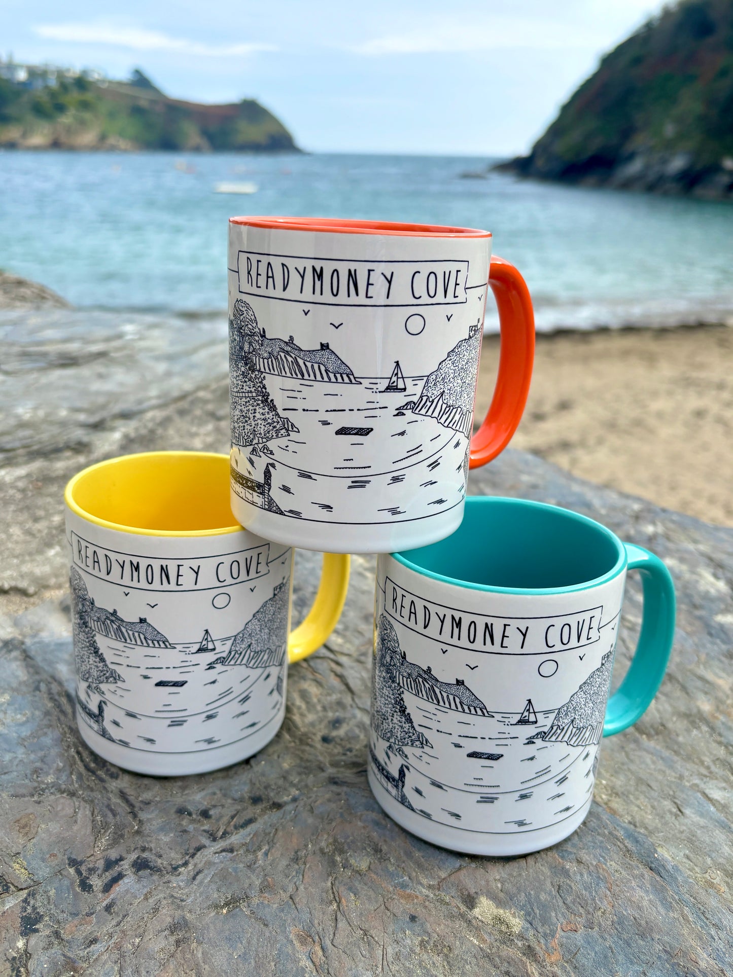 readymoney cove mugs with coastal illustration, colorful handles, beach backdrop—perfect seaside-themed gift or souvenir
