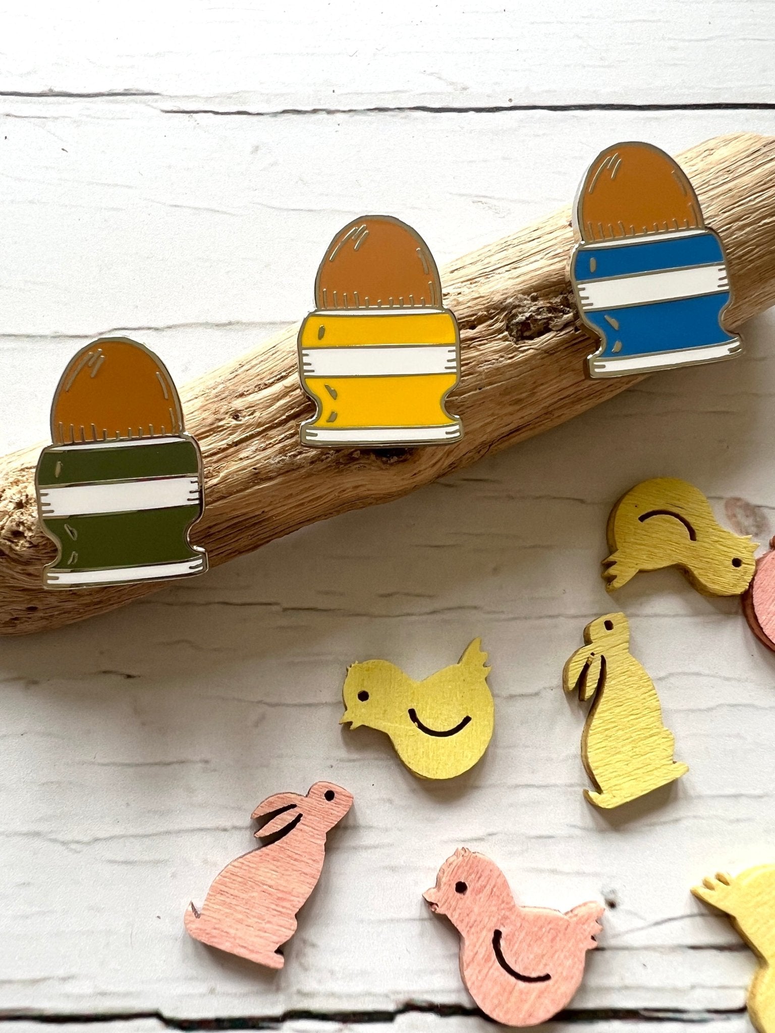 colorful striped easter egg enamel pin badges in egg cups on driftwood with wooden bunny and chick decorations