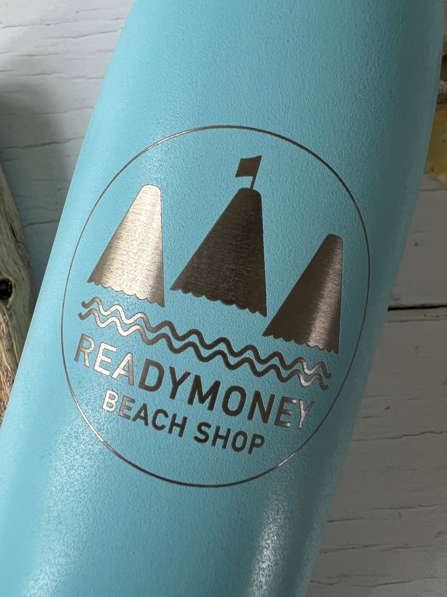 closeup shot of blue water bottle showing metallic silver Readymoney beach shop engraved logo with sailboat design