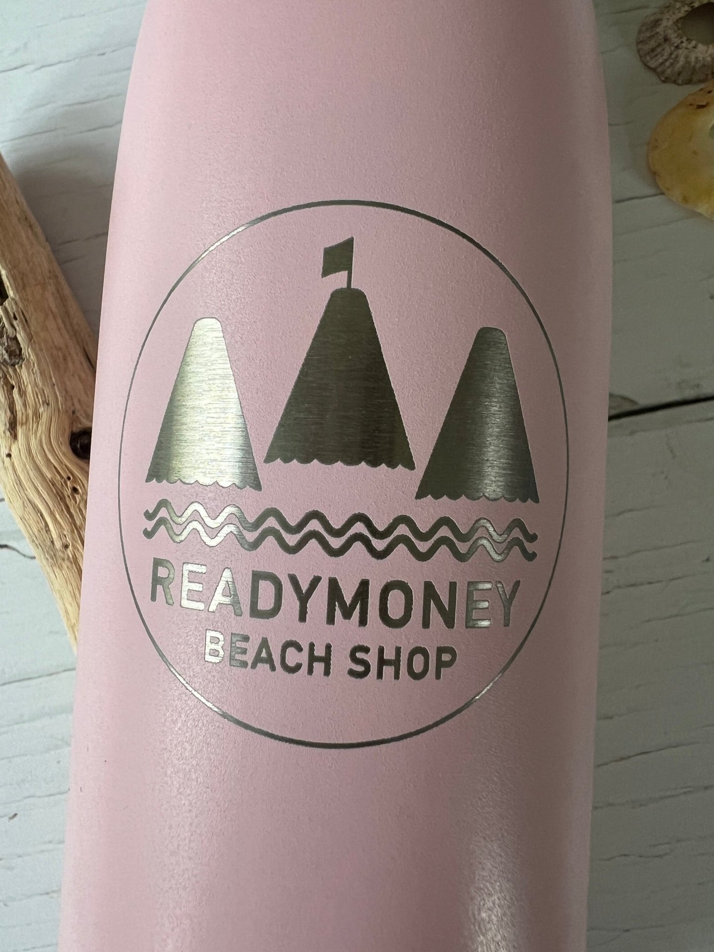 closeup shot of pink water bottle showing silver Readymoney beach shop engraved logo with sailboat and waves detail