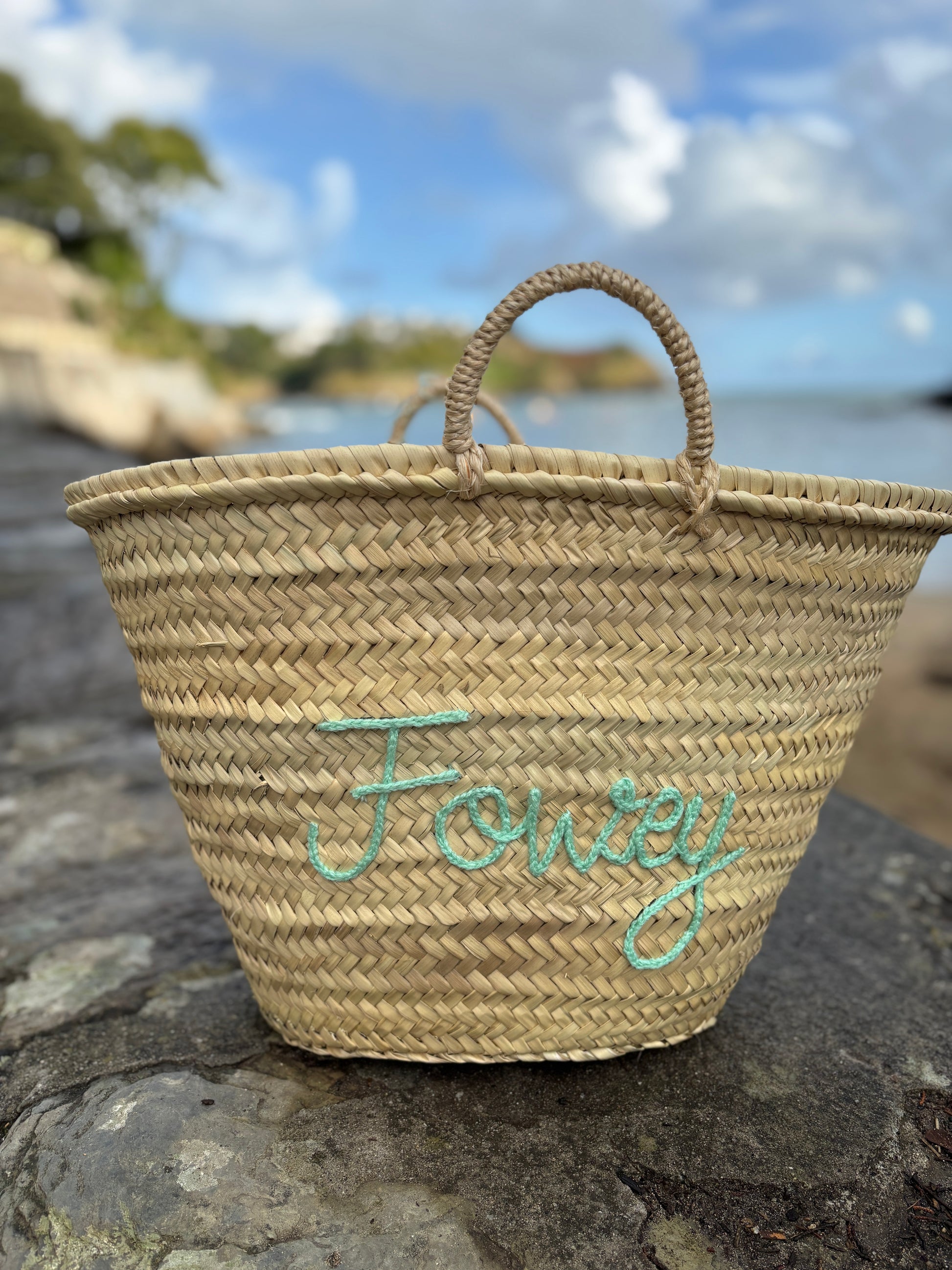 hand-woven straw basket bag with Fowey seafoam green embroidery customized beach tote or coastal holiday accessory photographed by the sea