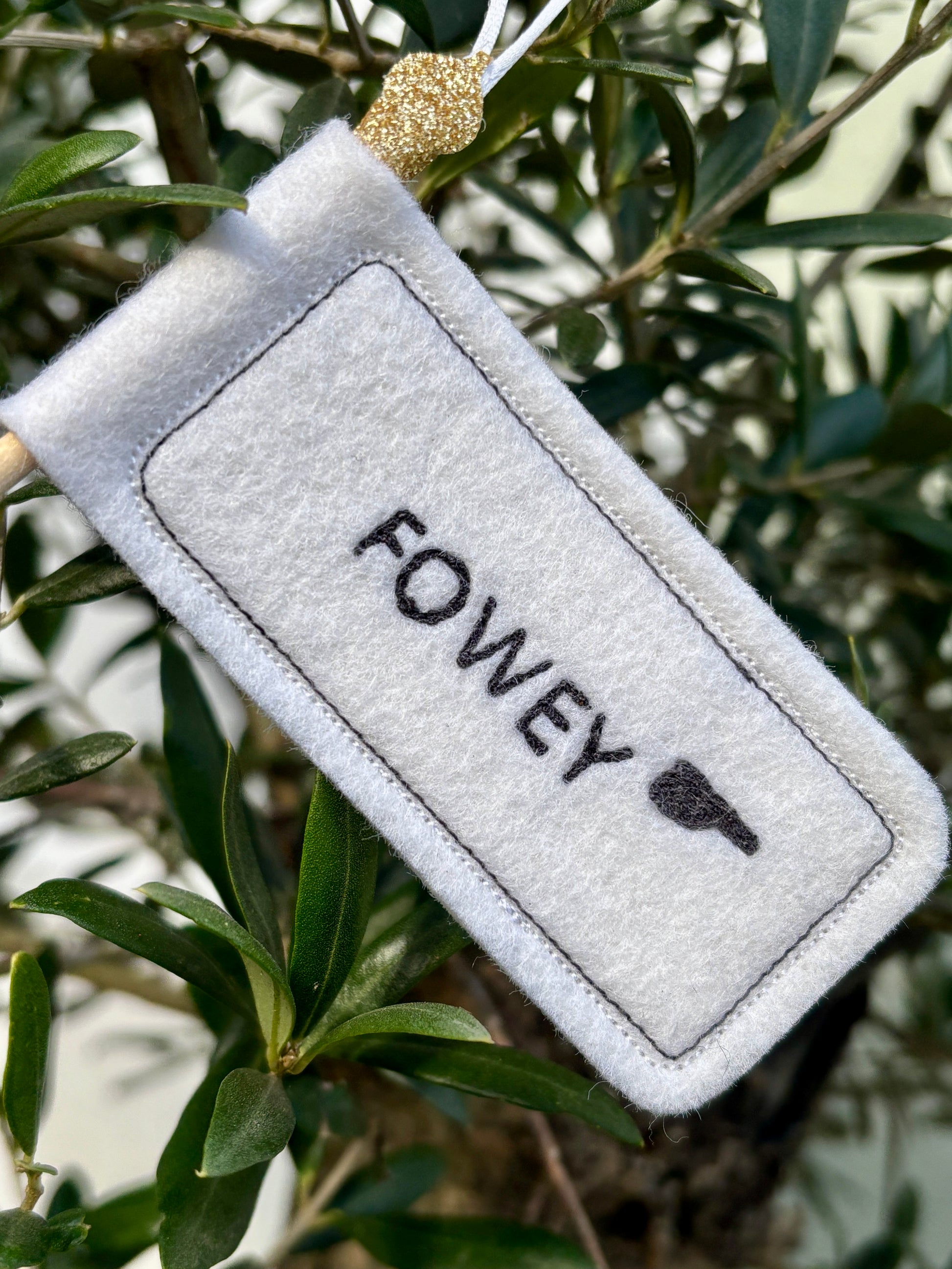 closeup shot of felt 'Fowey' signpost handmade white textile ornament with a glitter loop, resting on a green tree branch seaside town gift