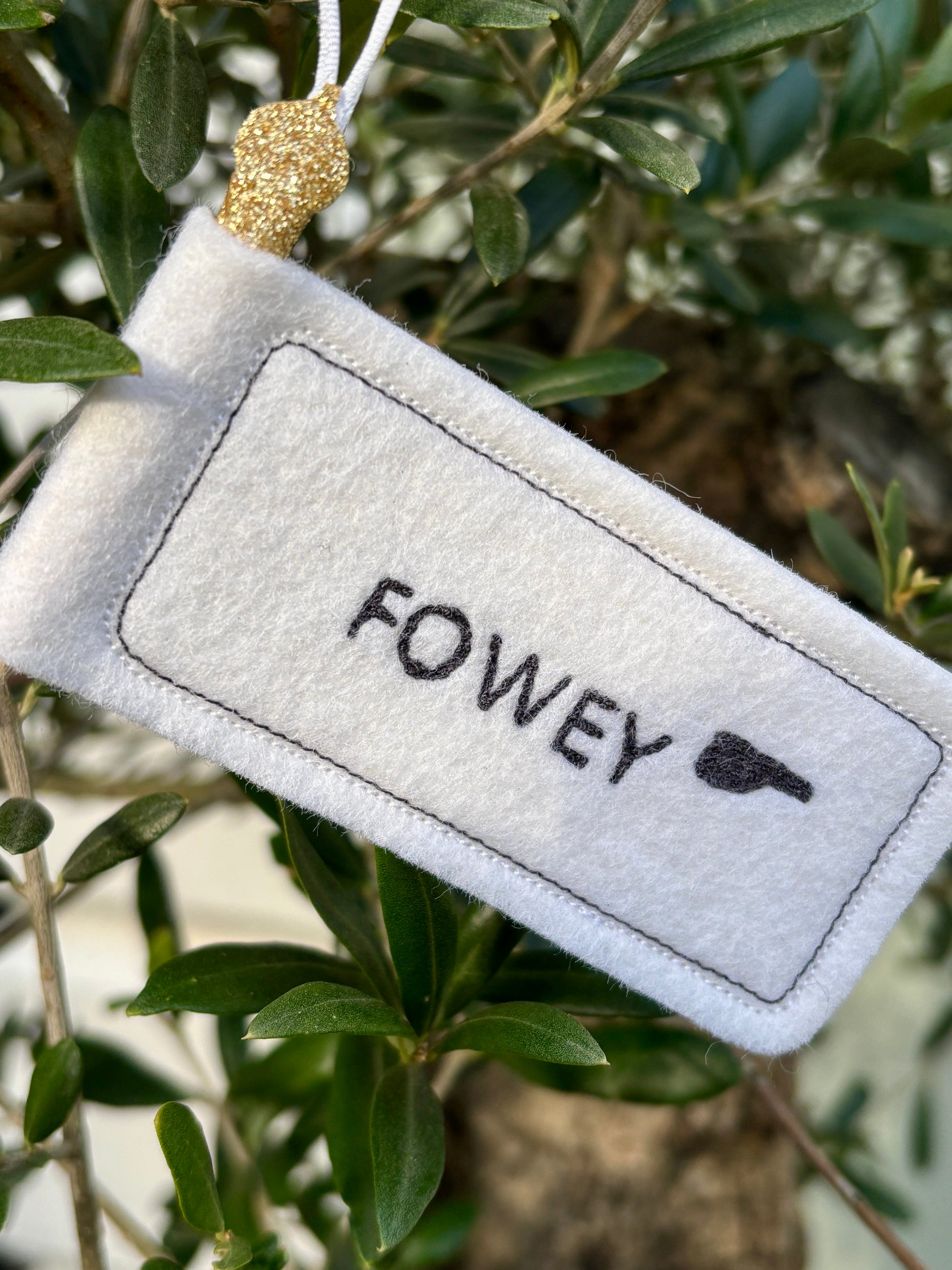 closeup shot of felt 'Fowey' signpost handmade white textile ornament with a glitter loop, resting on a green tree branch seaside town gift