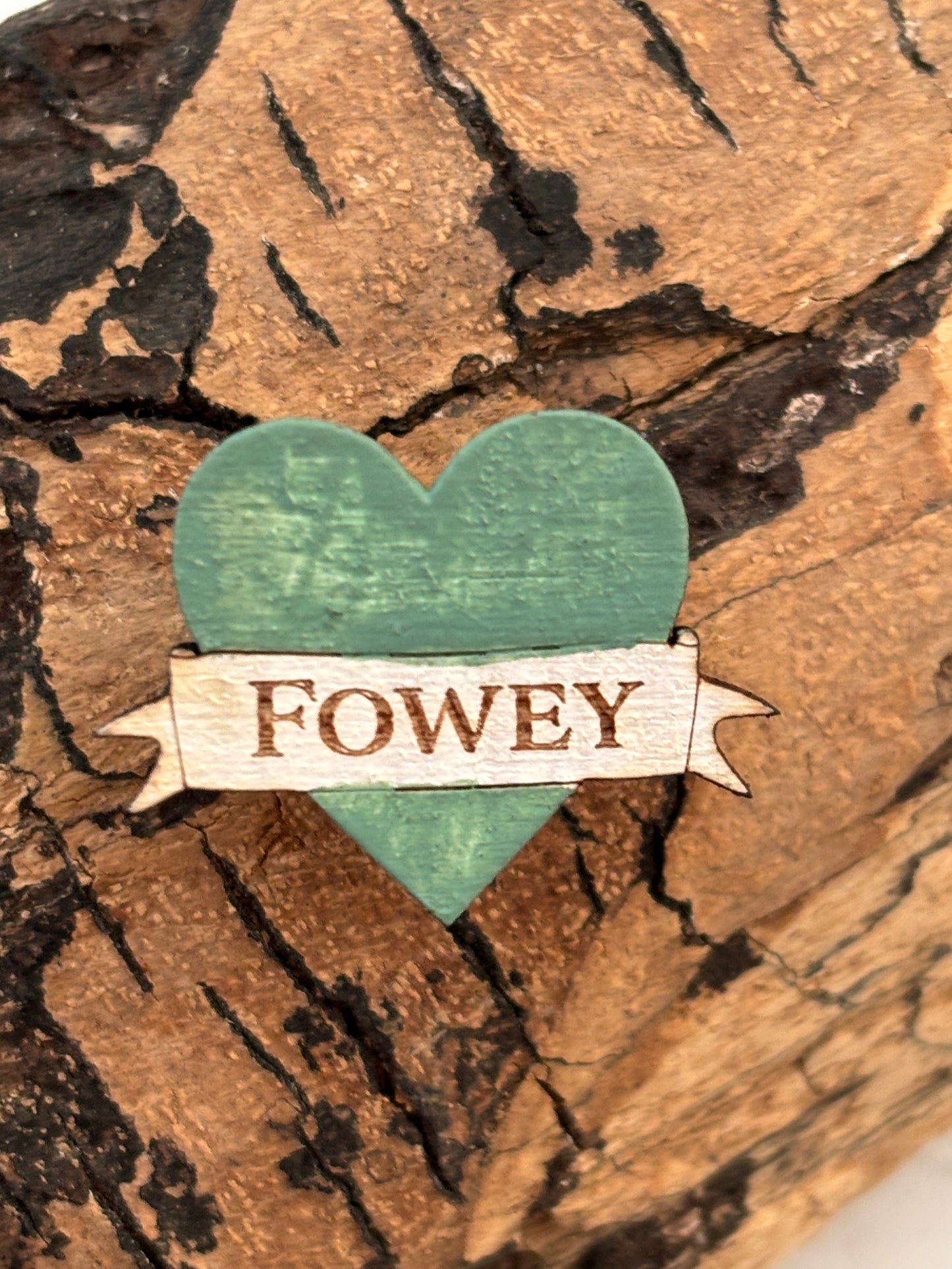 closeup shot of the Fowey wooden heart pin teal wooden heart badge with an engraved Fowey banner on a piece of driftwood coastal souvenir accessory