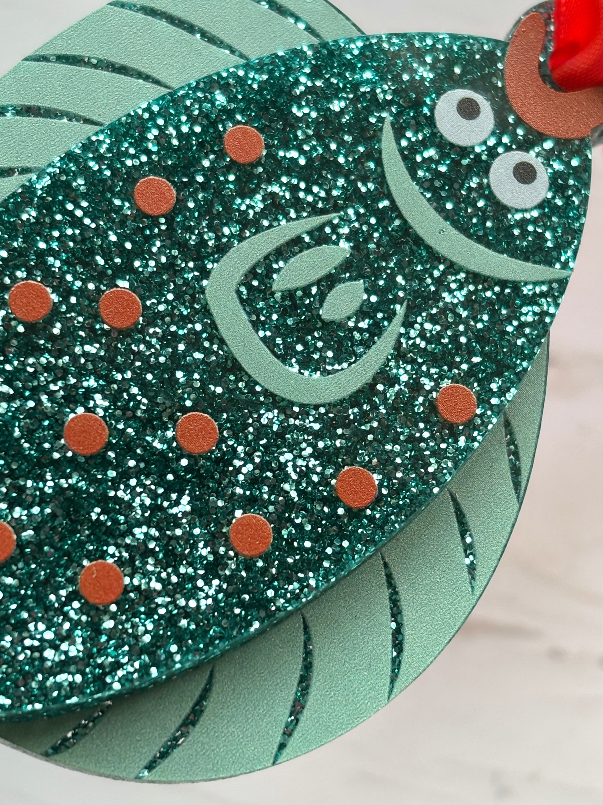 closeup shot of teal glittery plaice fish Christmas decoration with orange dots and red ribbon, fun coastal holiday ornament