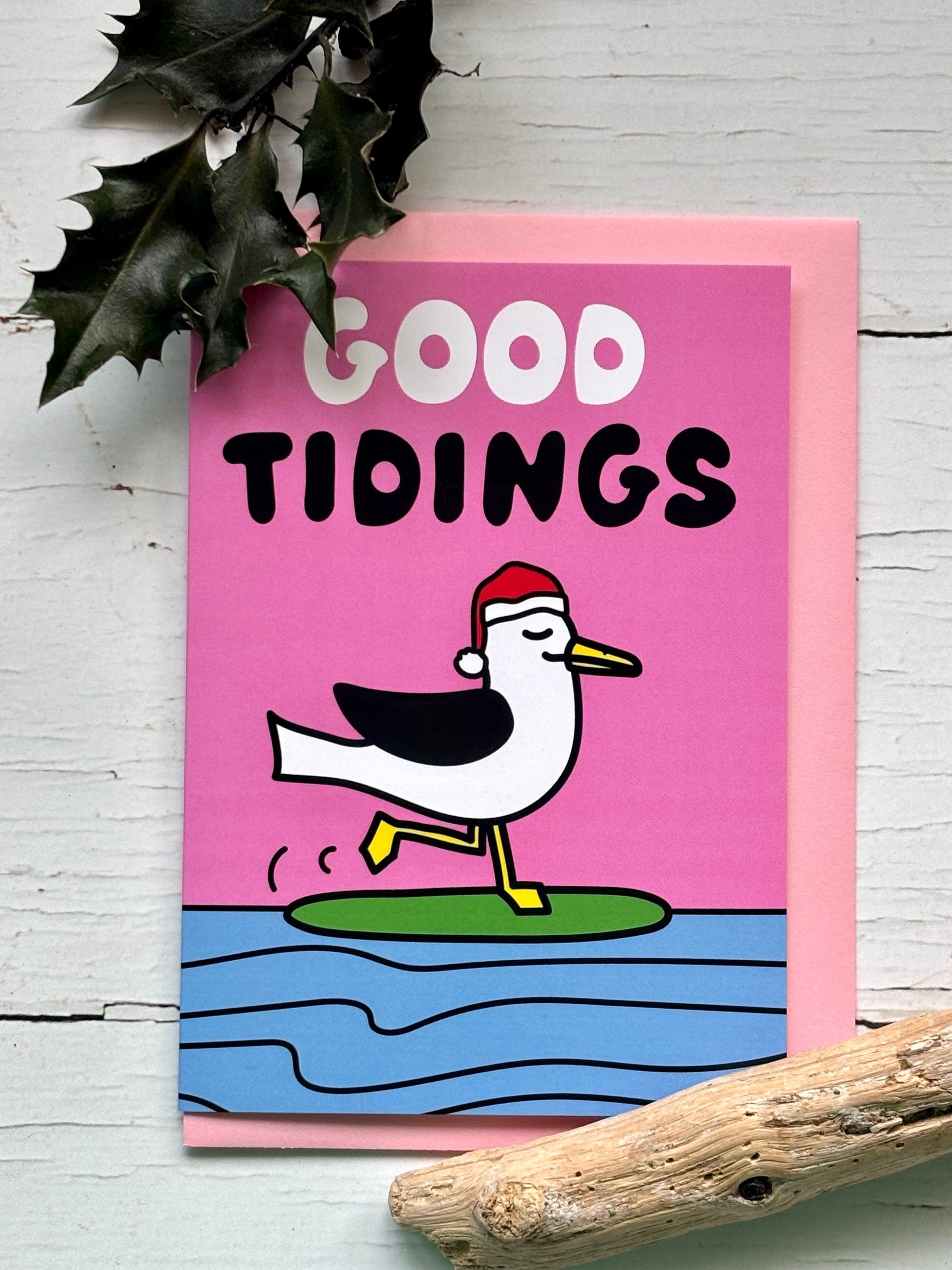 Cartoon seagull in Santa hat with “Good Tidings” text on pink background, standing happily on green surfboard