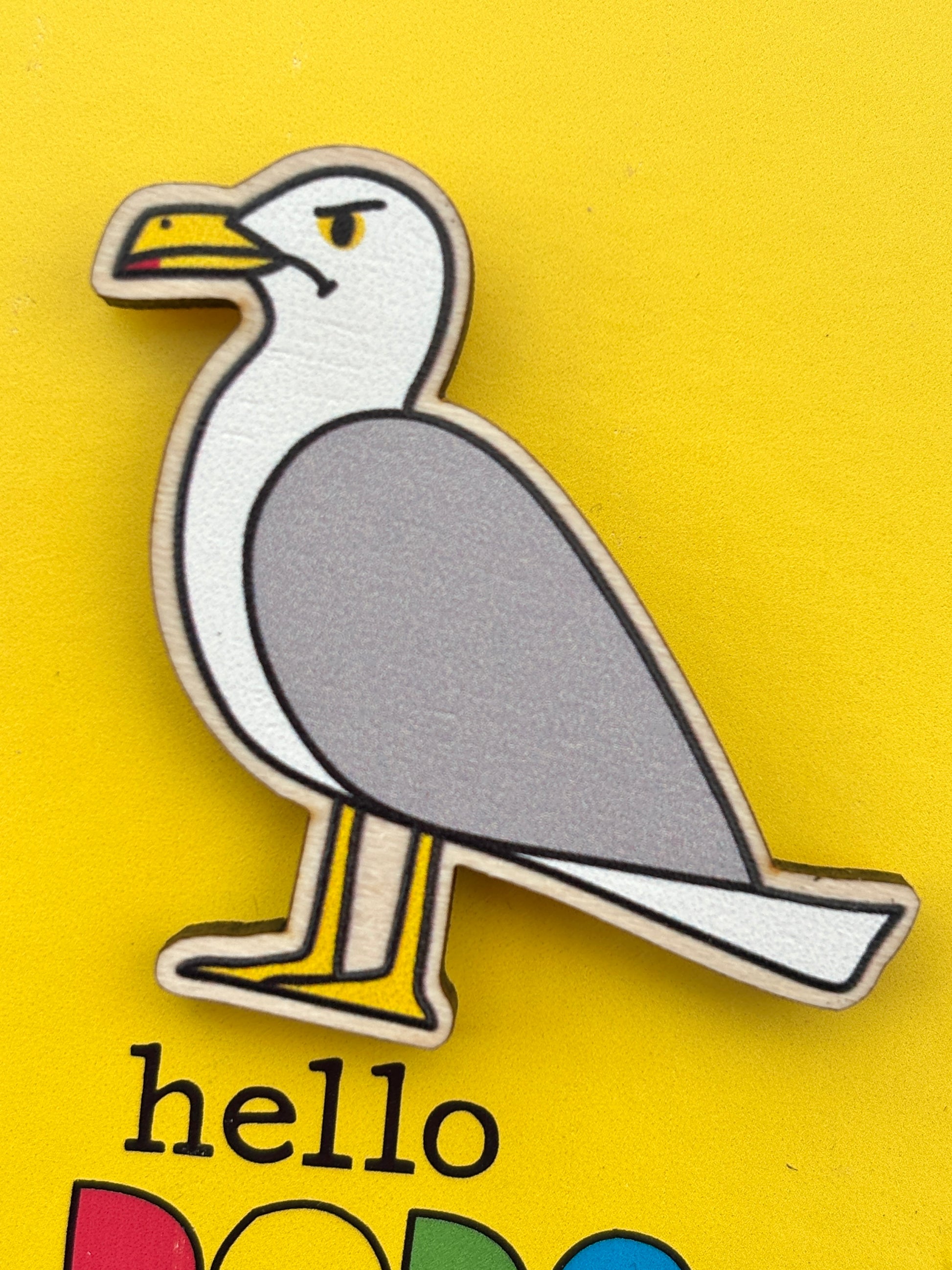 closeup shot of a grumpy seagull wooden pin badge on a yellow background funky nautical accessory, great eco-friendly gift for bird lovers