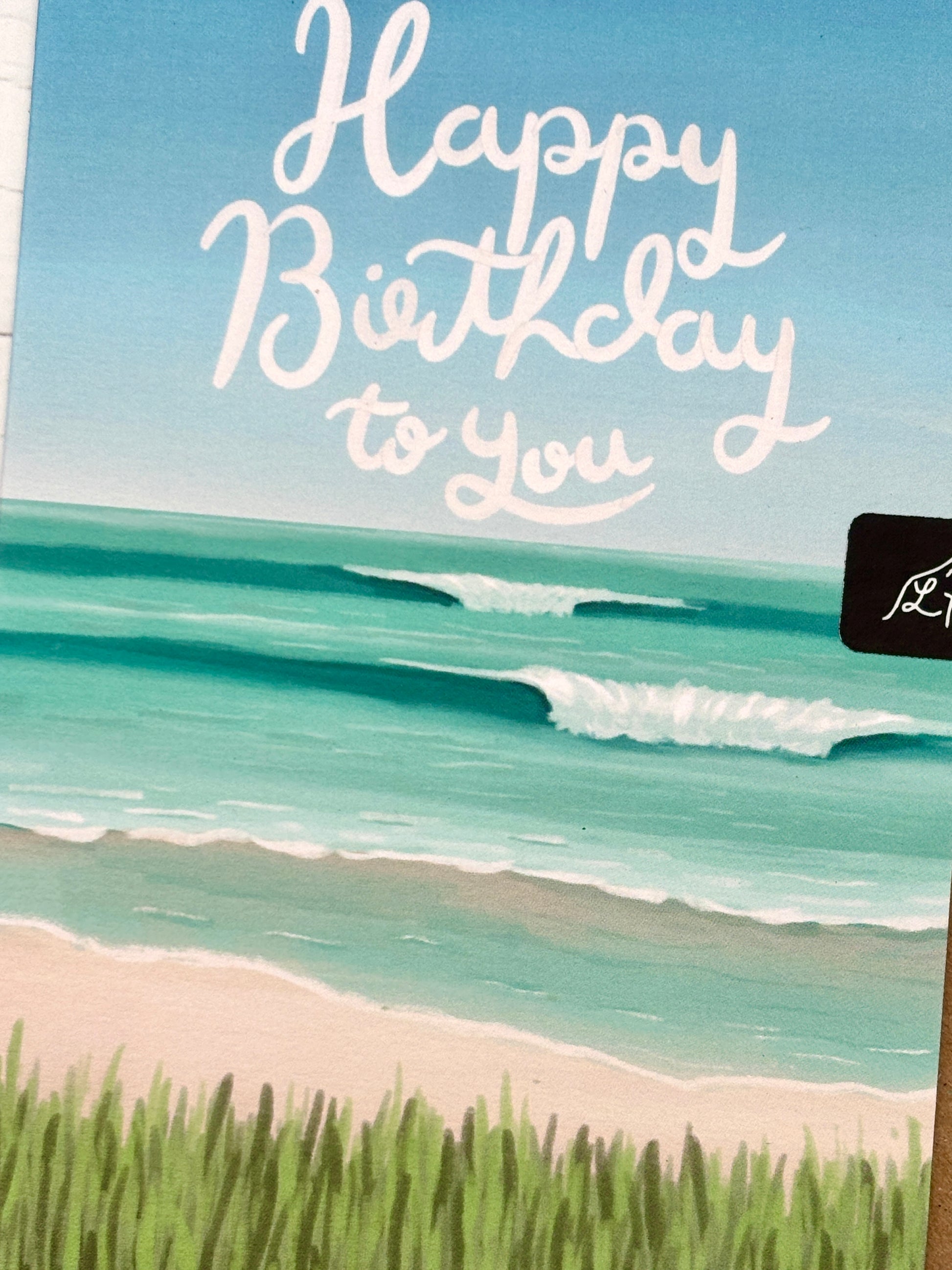 closeup shot of a Happy Birthday card showing a hand-painted turquoise ocean scene and beach grass, with hand-lettering above the waves coastal greeting card