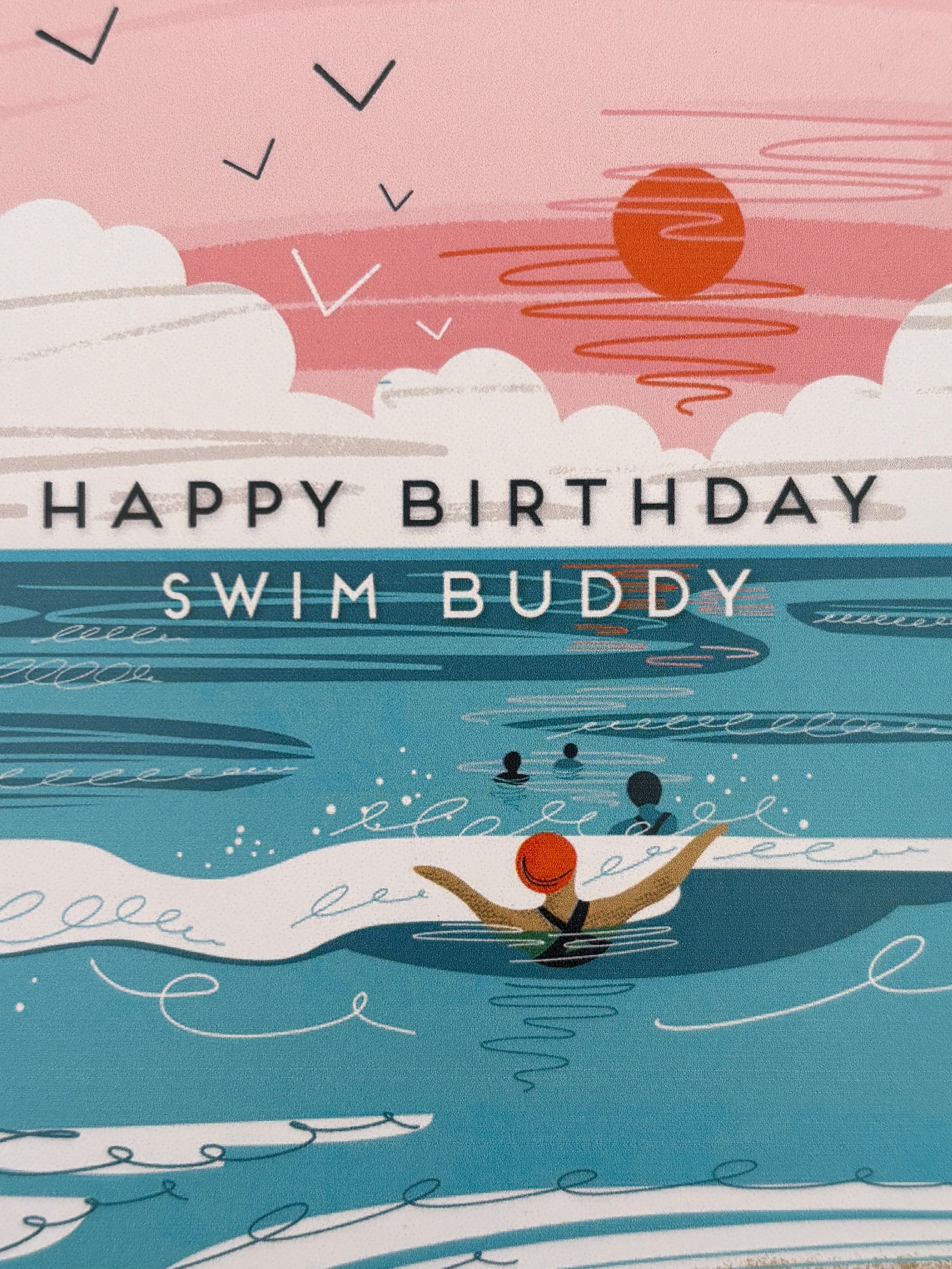 closeup shot of a Happy Birthday Swim Buddy card with an illustration of swimmers in the turquoise ocean and a sunset sky coastal greeting card for sea swimmers