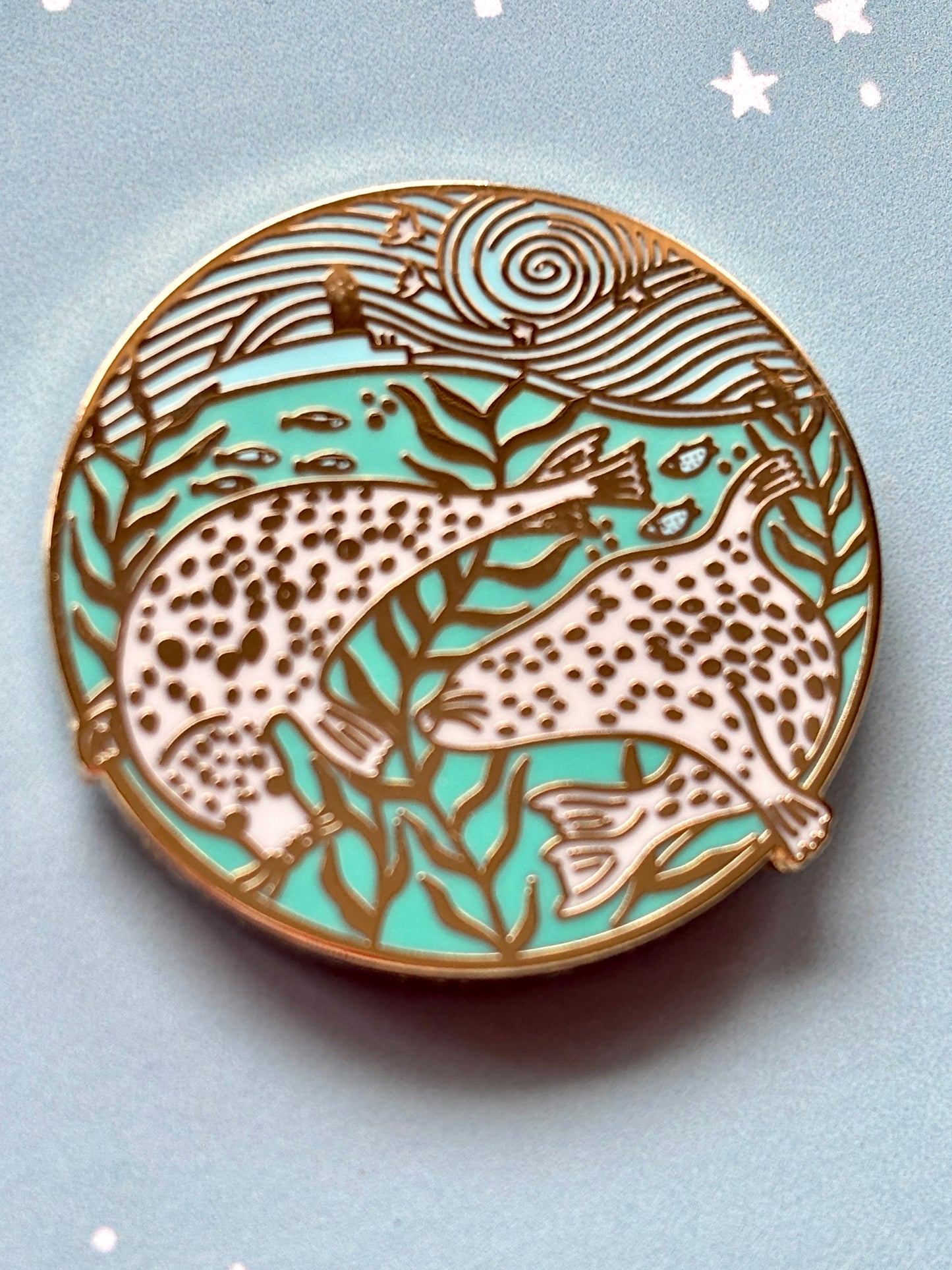 closeup shot of harbour seals enamel pin with gold reef, lighthouse, and swirling sea for marine life lovers and pin collectors