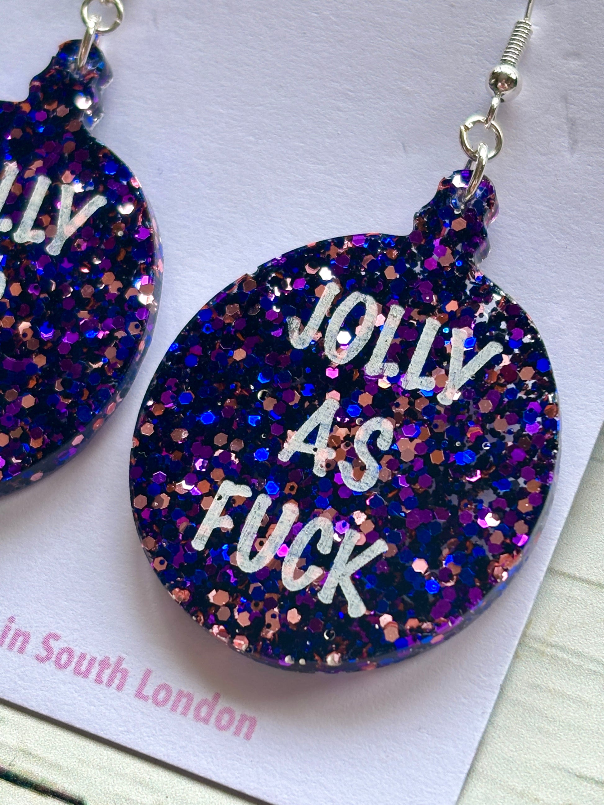 closeup shot of novelty acrylic earring glitter bauble shape with bold, humorous Christmas text unique adult holiday accessory