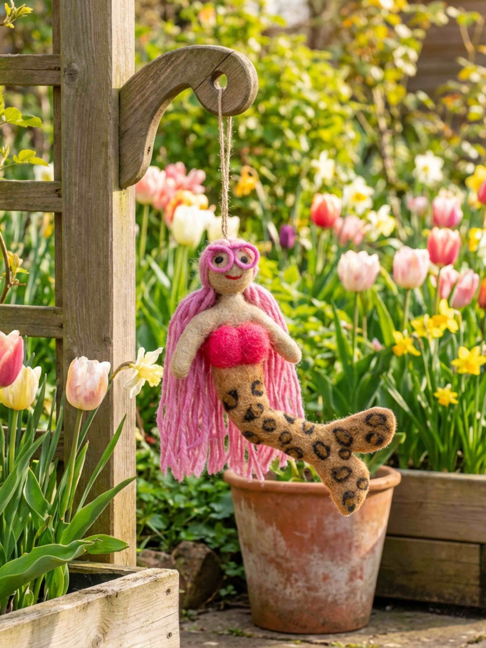 A handmade felt mermaid hanging from a wooden garden trellis, with a pink yarn tail, pink top, and leopard-print fin, set against a blurred background of a garden with tulips and flowers.