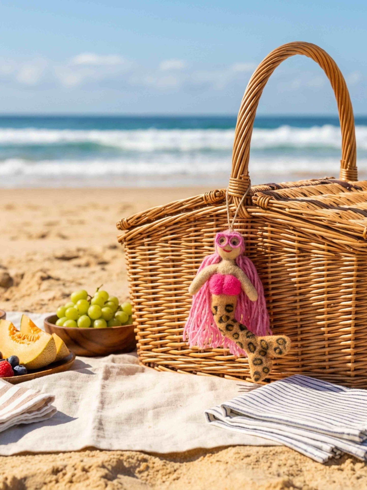 A wicker picnic basket on a sandy beach with the ocean in the background. A small mermaid with pink hair and sunglasses hangs from the basket handle, and a striped towel, grapes, and sliced melon are arranged nearby.