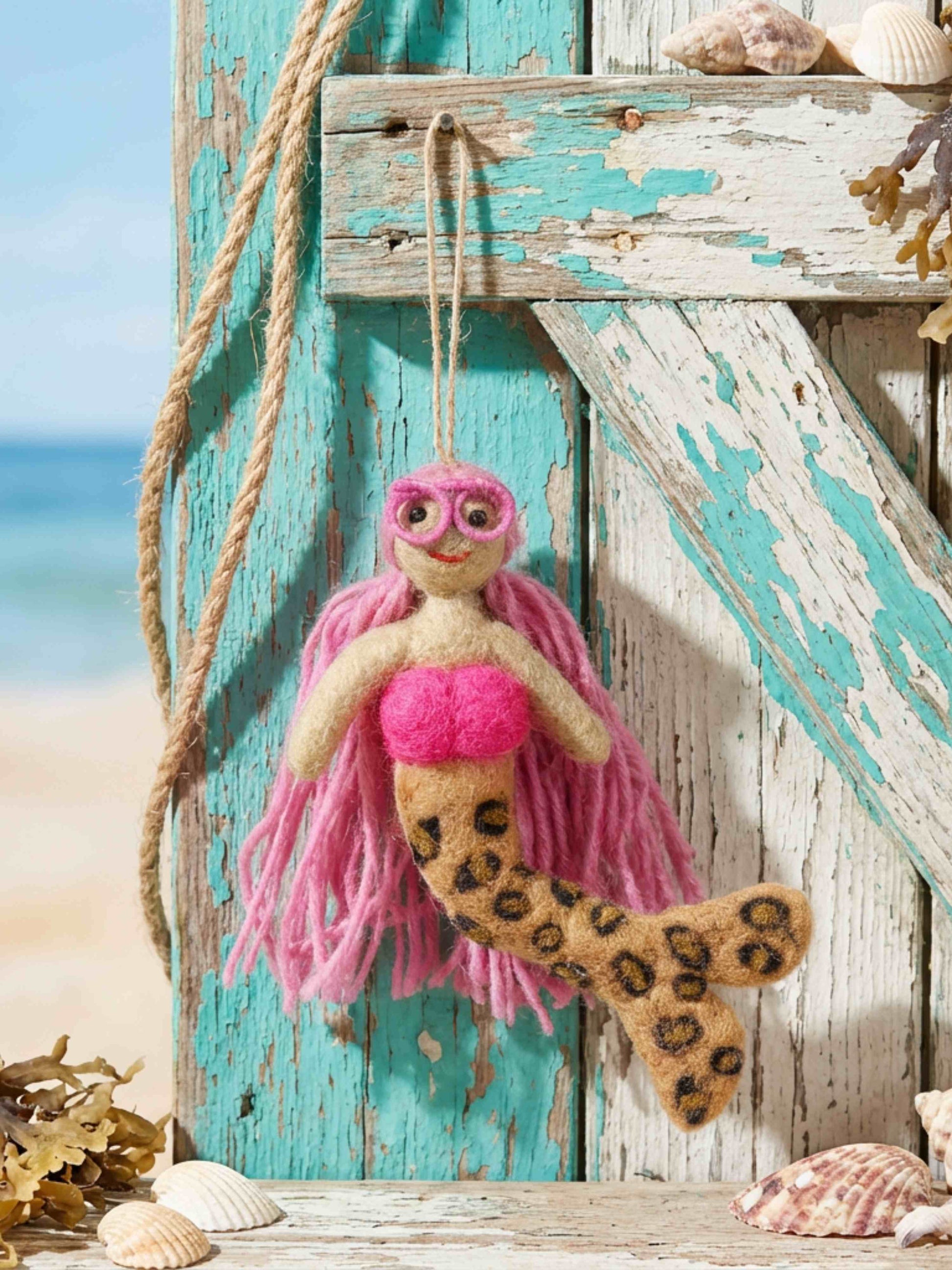 A handmade felt mermaid ornament hanging on a rustic turquoise wooden door with seashells and rope nearby, against a blurred beach background.
