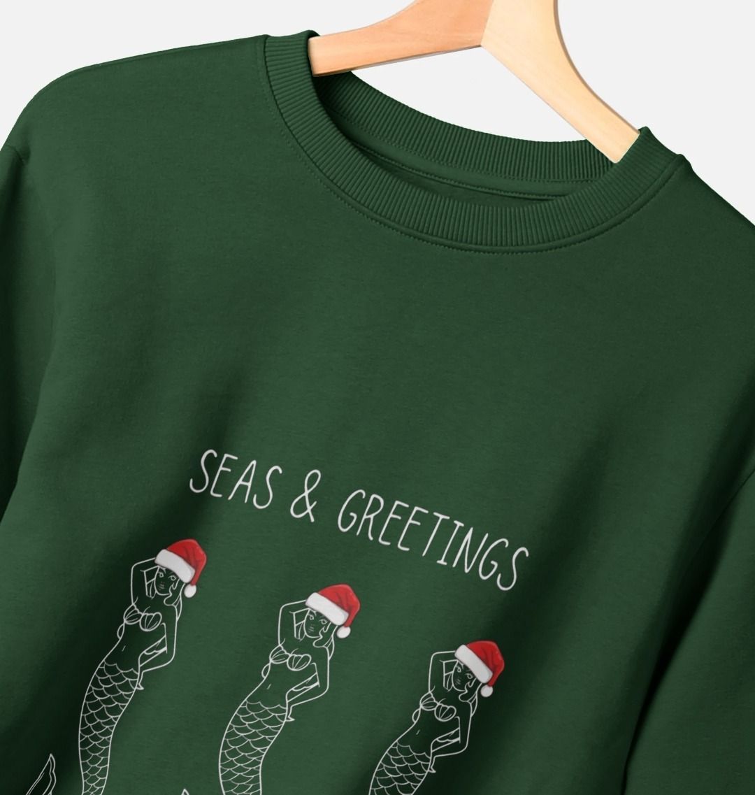 closeup shot of evergreen Christmas jumper with "Seas & Greetings" text features three mermaids wearing Santa hats festive, funny apparel