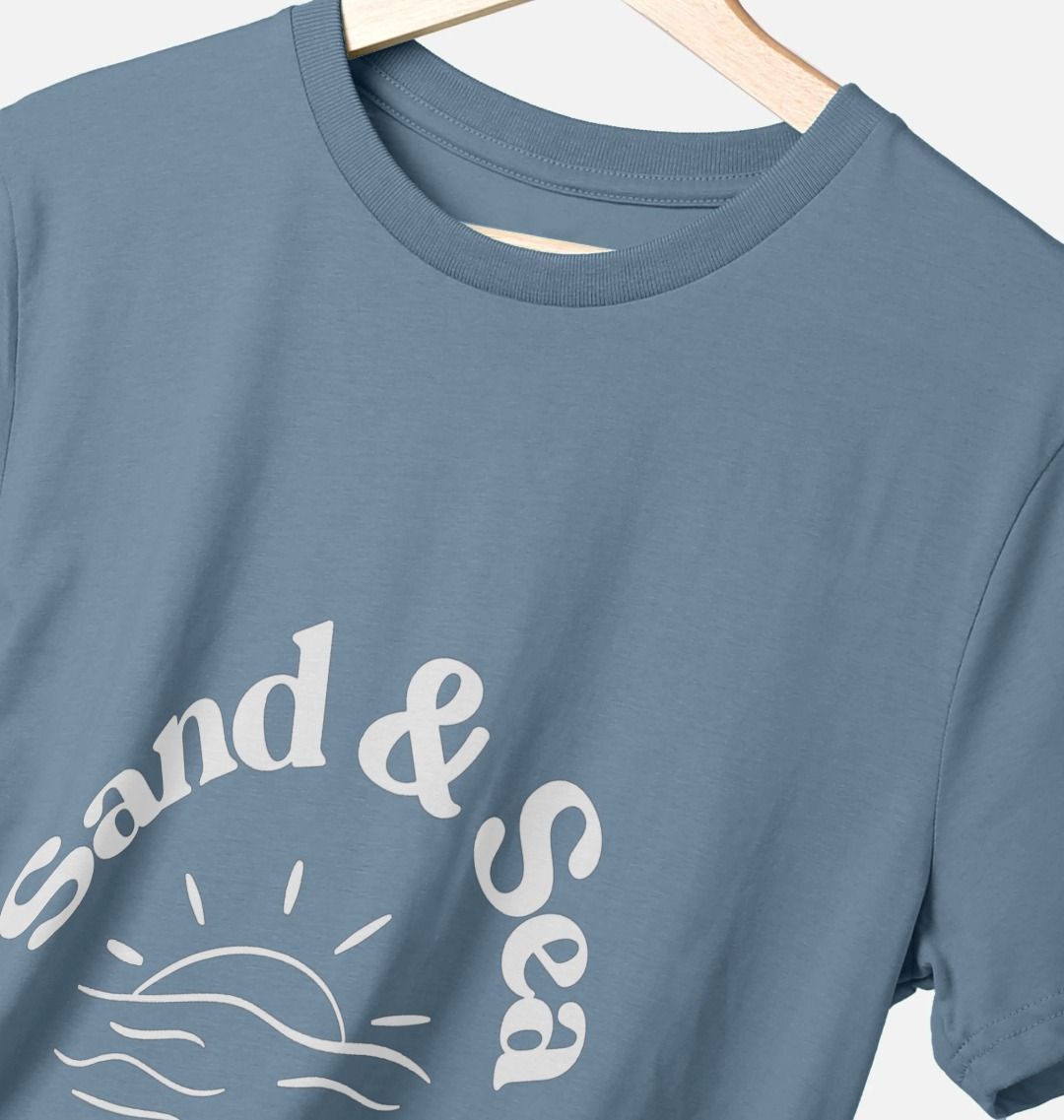 closeup shot of stone blue T-shirt with white "Sand & Sea" graphic features sun and wave detail men's casual coastal apparel and eco-friendly tee