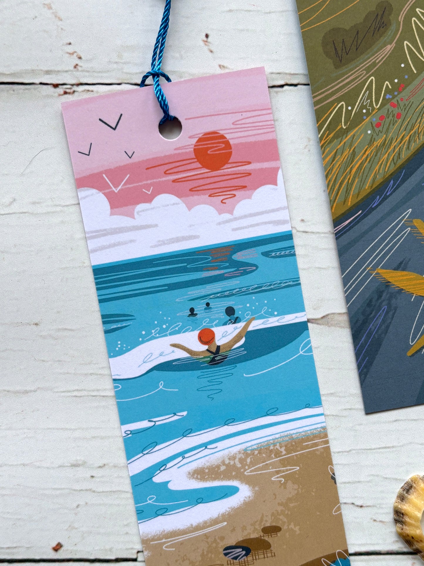 illustrated coastal bookmark wild swimming at the beach with a pink sunset and blue tassel seaside reader gift