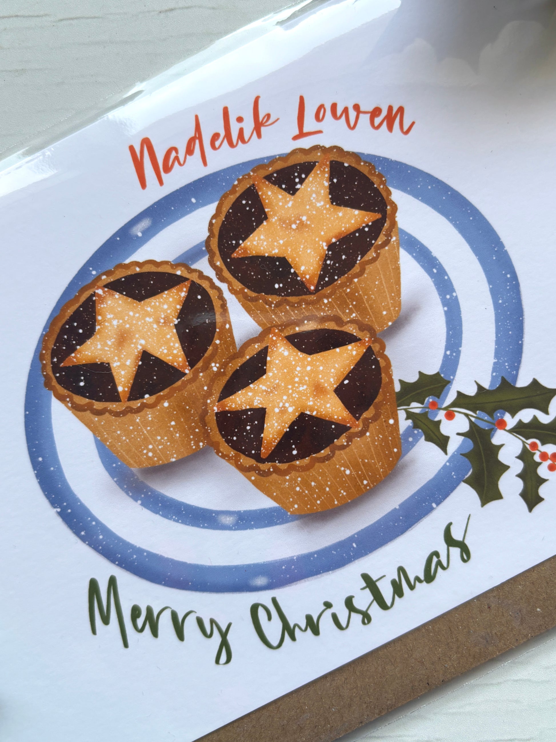 closeup shot of Cornish Christmas card with star-topped mince pies, powdered sugar, holly sprig, and festive “Nadelik Lowen” greeting