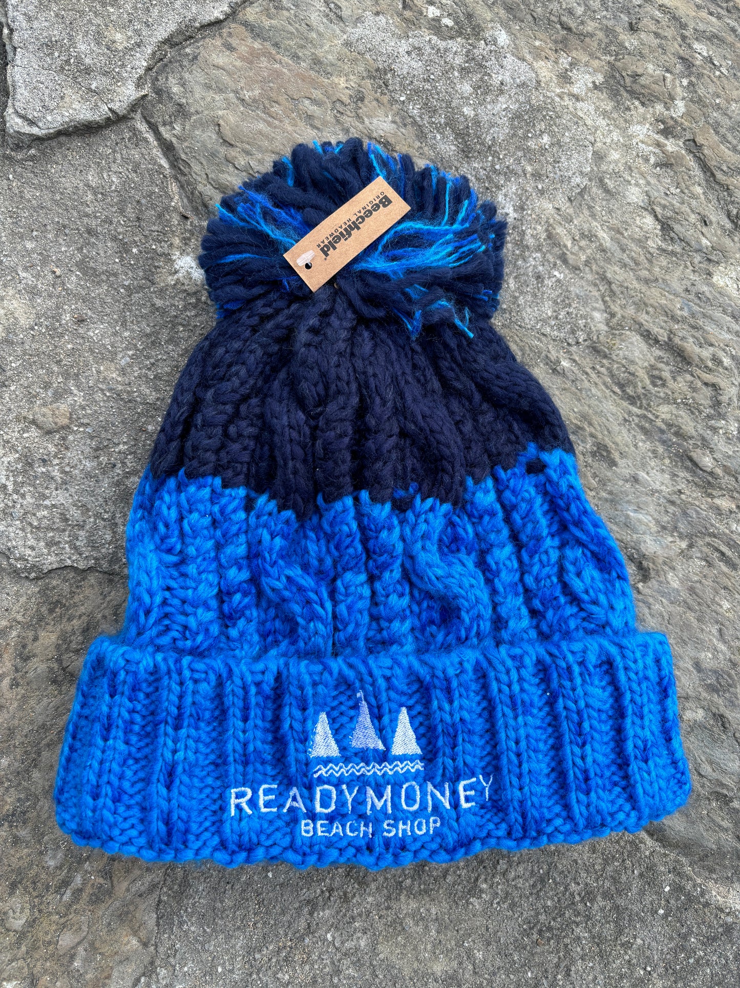blue-navy chunky bobble hat with faux fur pom-pom and beach shop logo—cozy, bold, and perfect for seaside winters