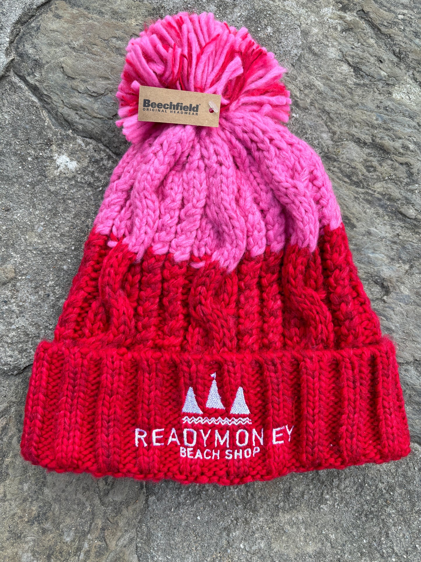 red-pink chunky bobble hat with faux fur pom-pom and beach shop logo—warm, bold, and winter-ready for kids