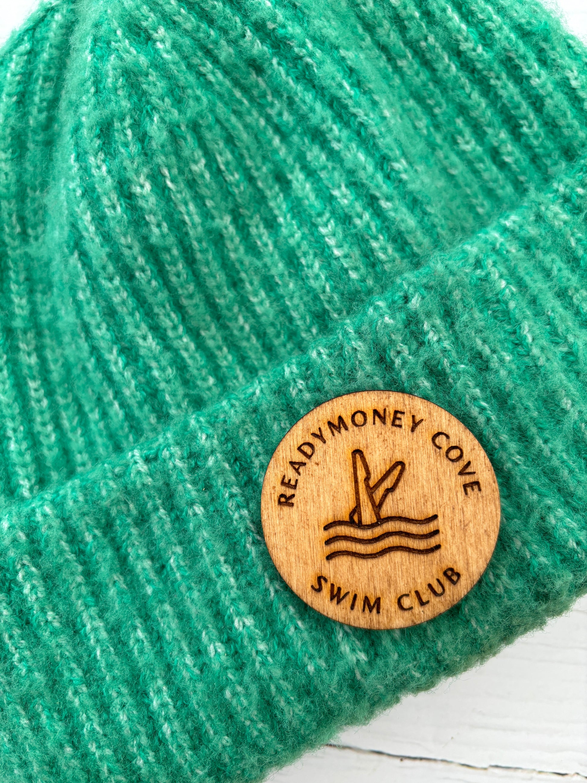 wooden 'Readymoney Cove Swim Club' circular badge pin worn on a green knitted beanie nautical, coastal souvenir, winter hat accessory