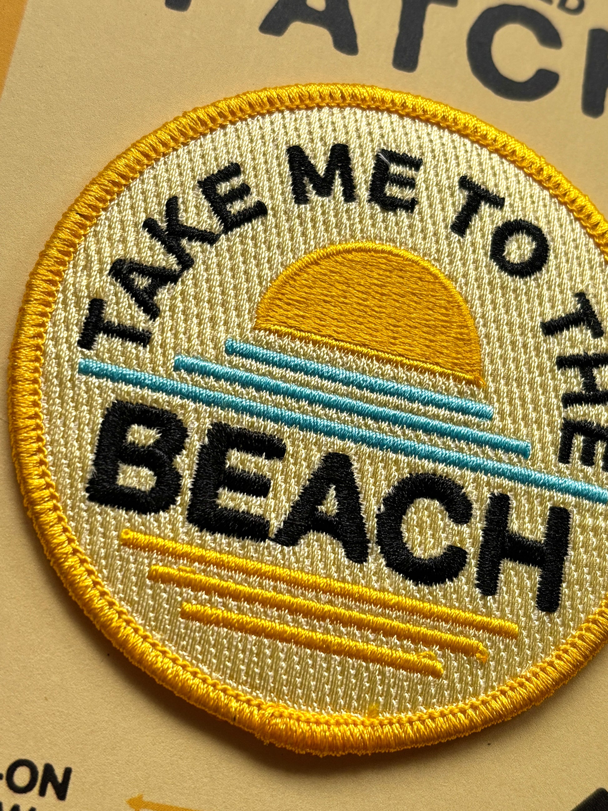 close up of a yellow fabric patch with a sun on and the words take me to the beach