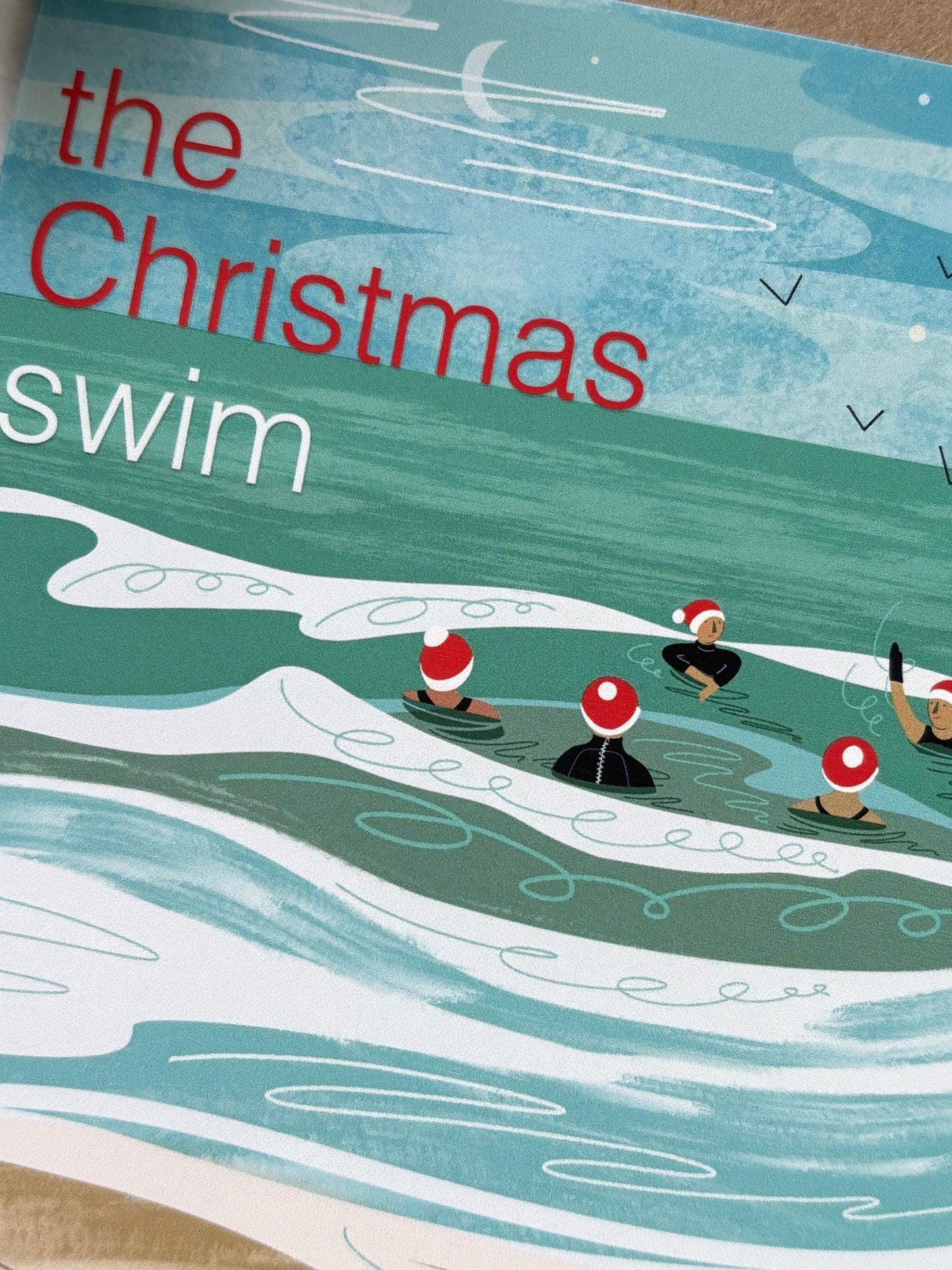 “The Christmas Swim” card close-up with swimmers in Santa hats in teal ocean waves, festive coastal holiday artwork