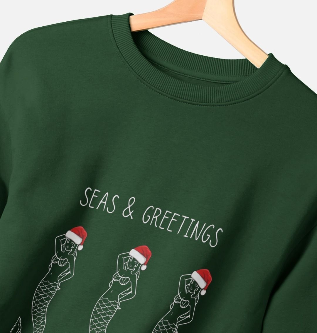 closeup shot of women's evergreen Christmas sweatshirt—outlined mermaids in Santa hats, “Seas & Greetings” festive graphic