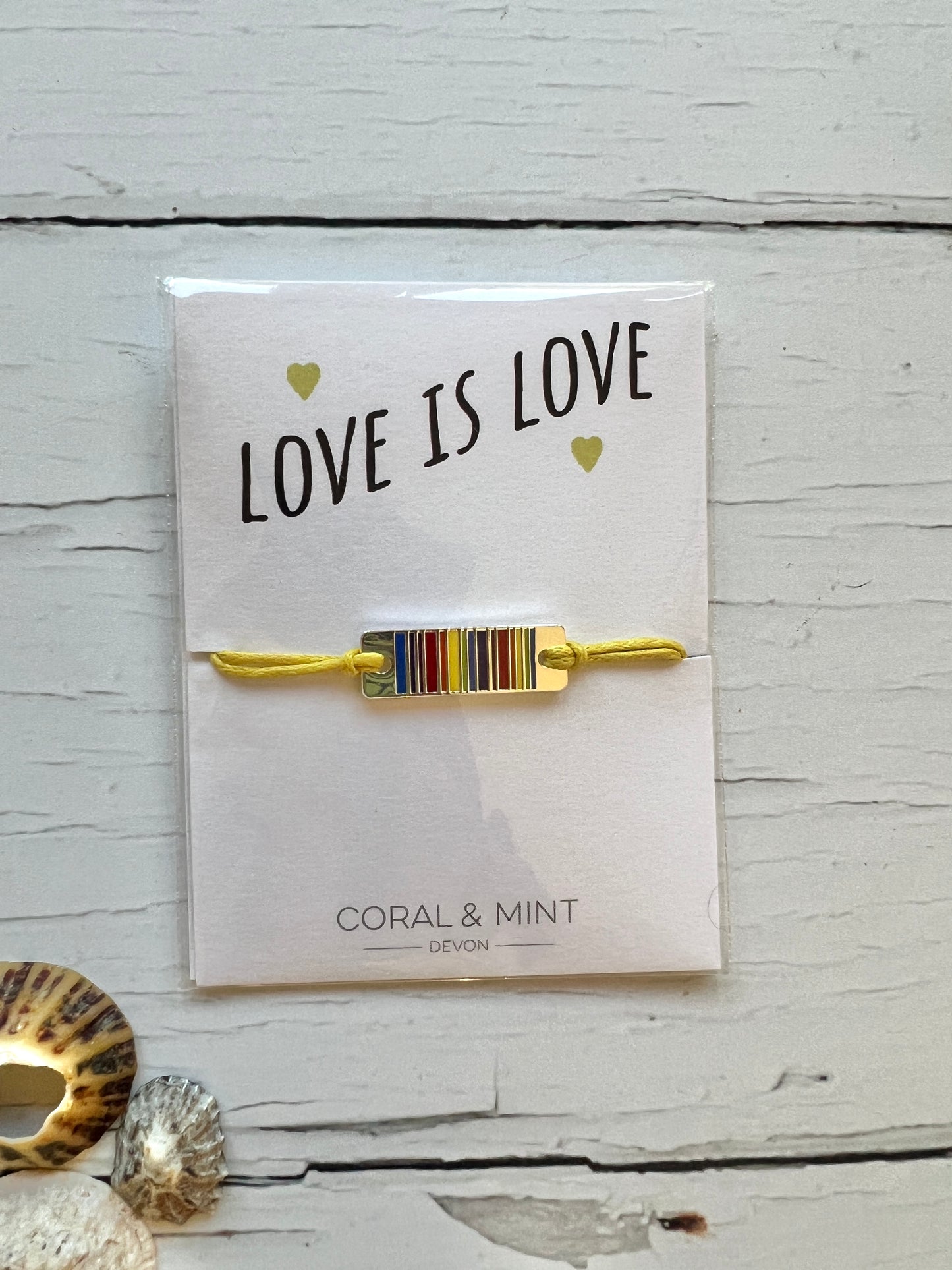 A string bracelet on a yellow cord with a silver rainbow striped charm. The text on the card reads Love is Love