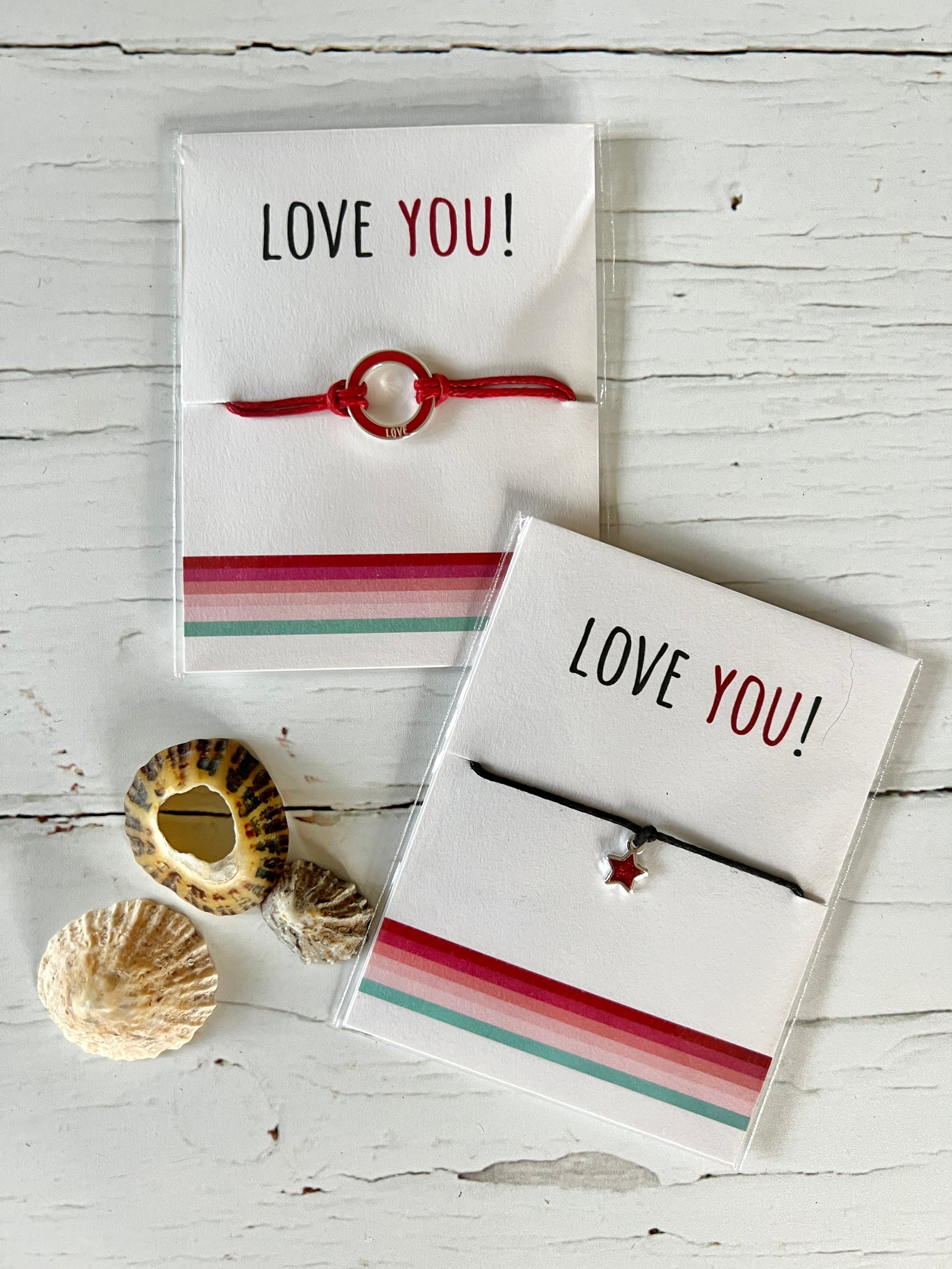 Two Valentine's string bracelets on presentation cards which say Love You. One bracelet has red cord and a red enamel disc and the other has a black cord and a red glittery enamel disc